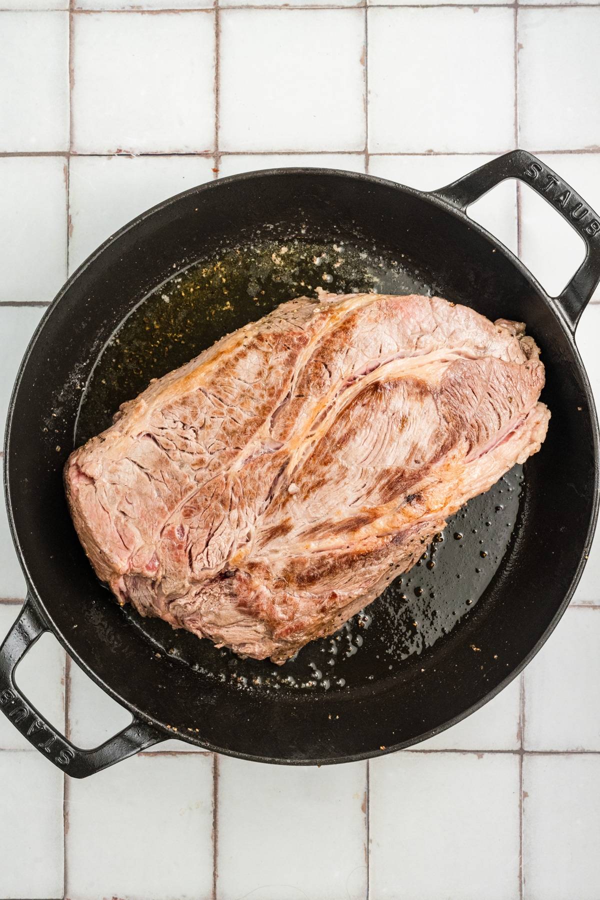 A large piece of seared beef in a black cast iron pot on a white tiled surface.