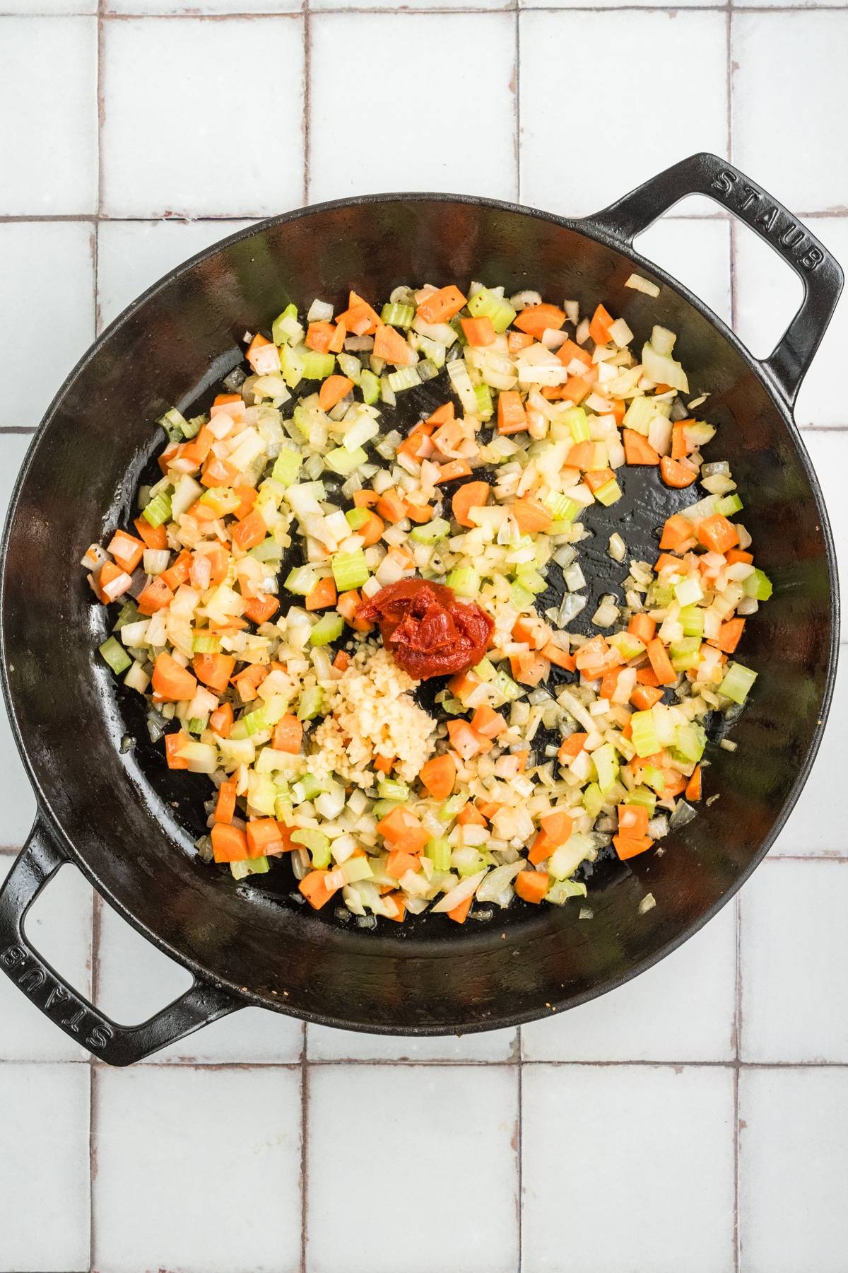 Diced carrots, celery, onion, garlic, and tomato paste in a black skillet on a tiled surface.