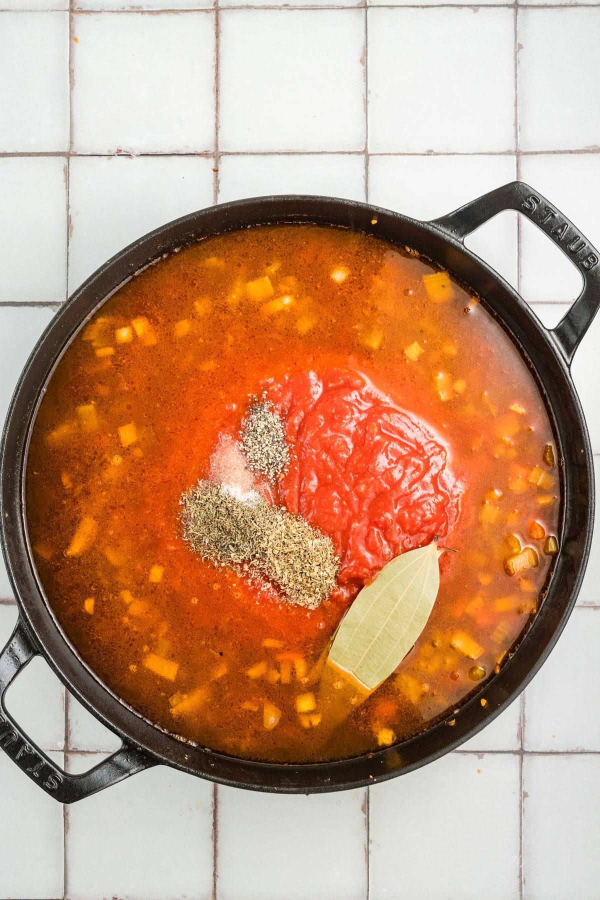 Black pot of soup with diced vegetables, tomatoes, herbs, and a bay leaf on a light tiled surface.