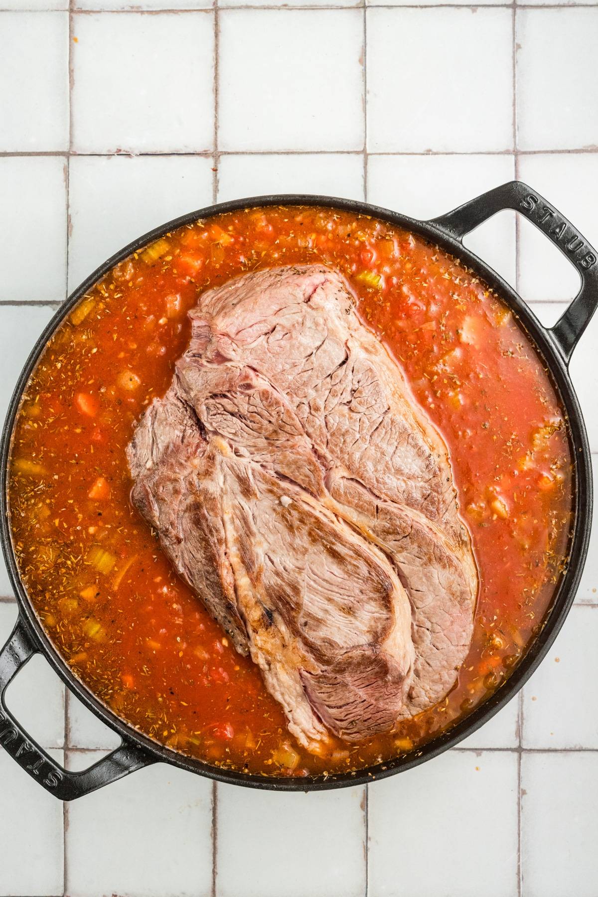 A large cut of beef in a pot of tomato sauce with vegetables on a tiled surface.