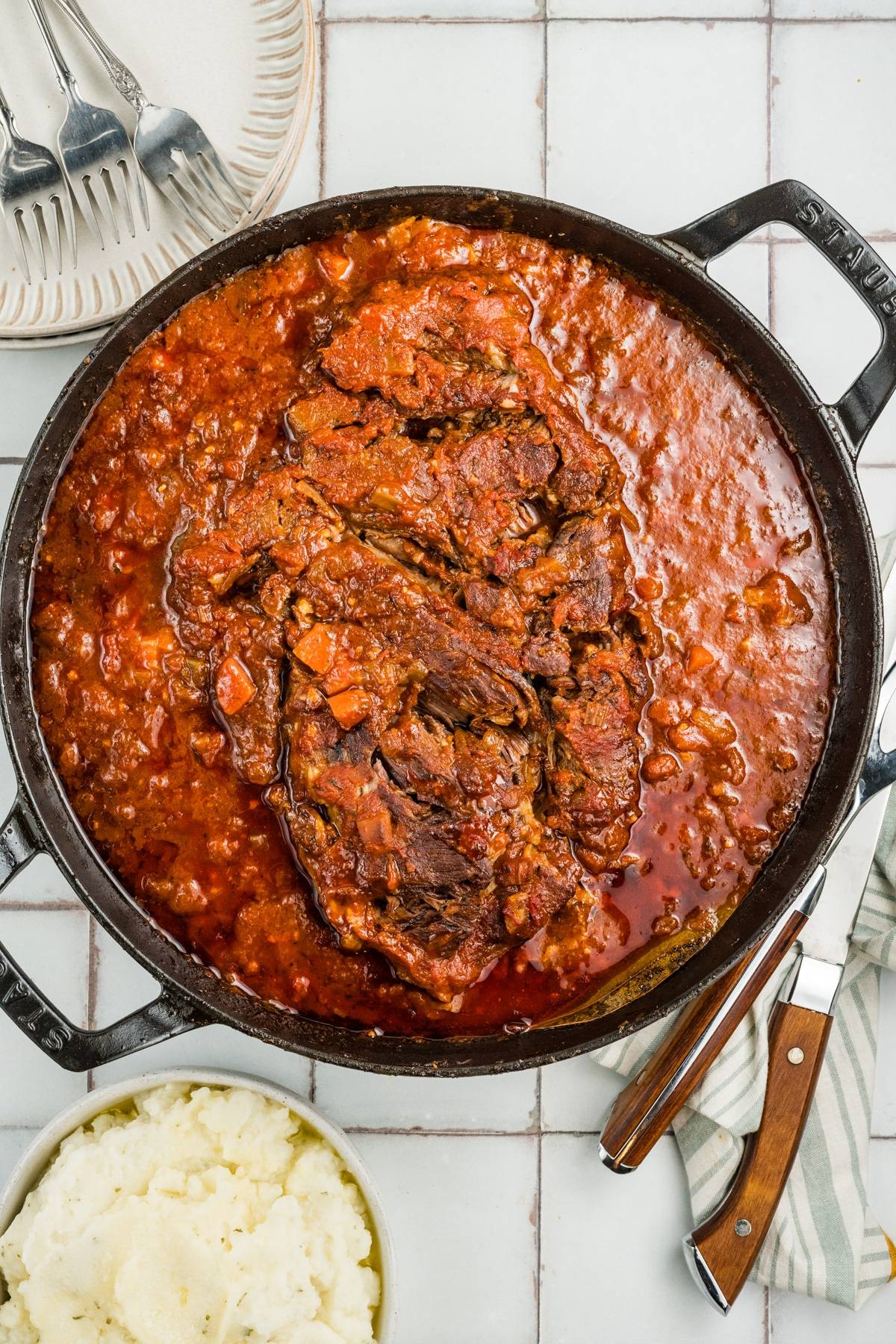 Large pot of braised beef in tomato sauce, accompanied by mashed potatoes, plates, and cutlery on the side.