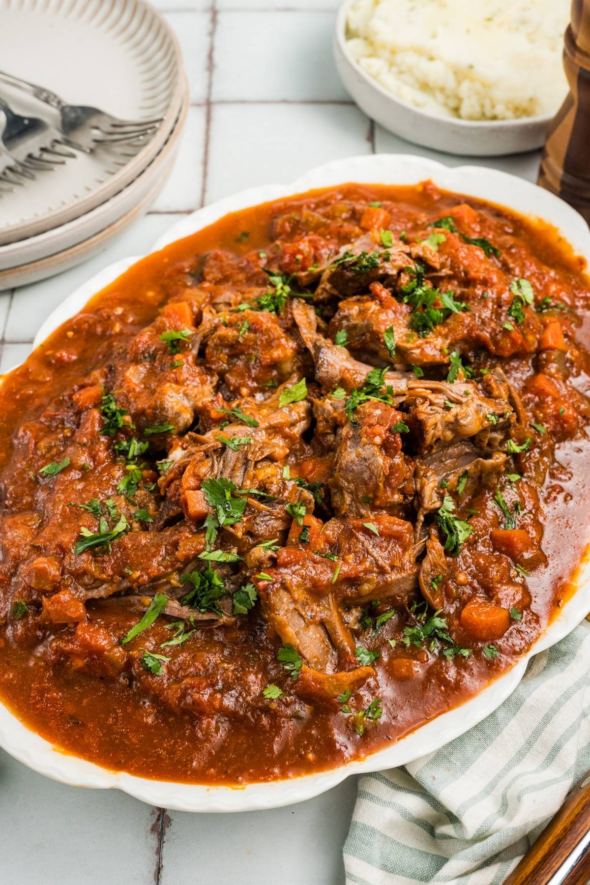 A platter of shredded beef in tomato sauce, garnished with chopped herbs, next to mashed potatoes.