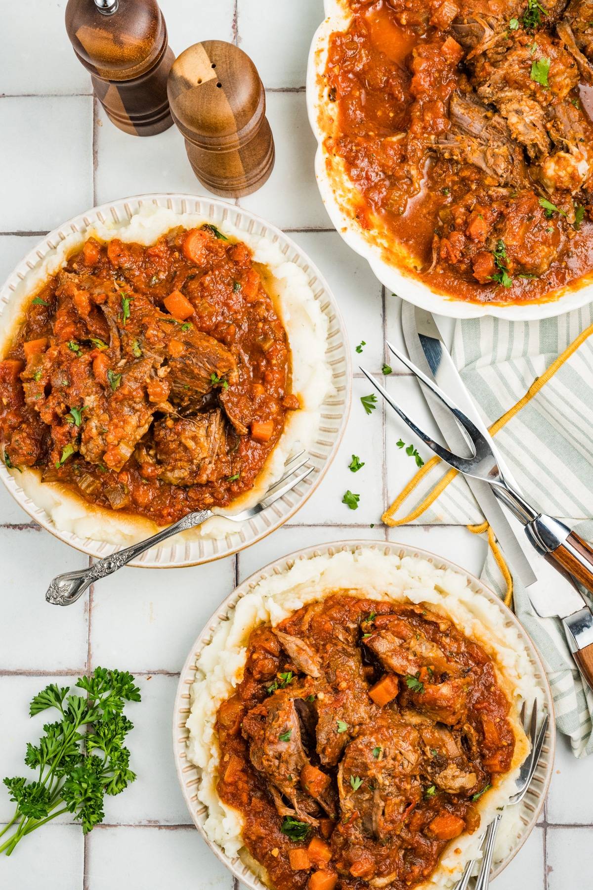 Overhead view of plates with pot roast in tomato sauce, garnished with parsley, on a tiled surface.
