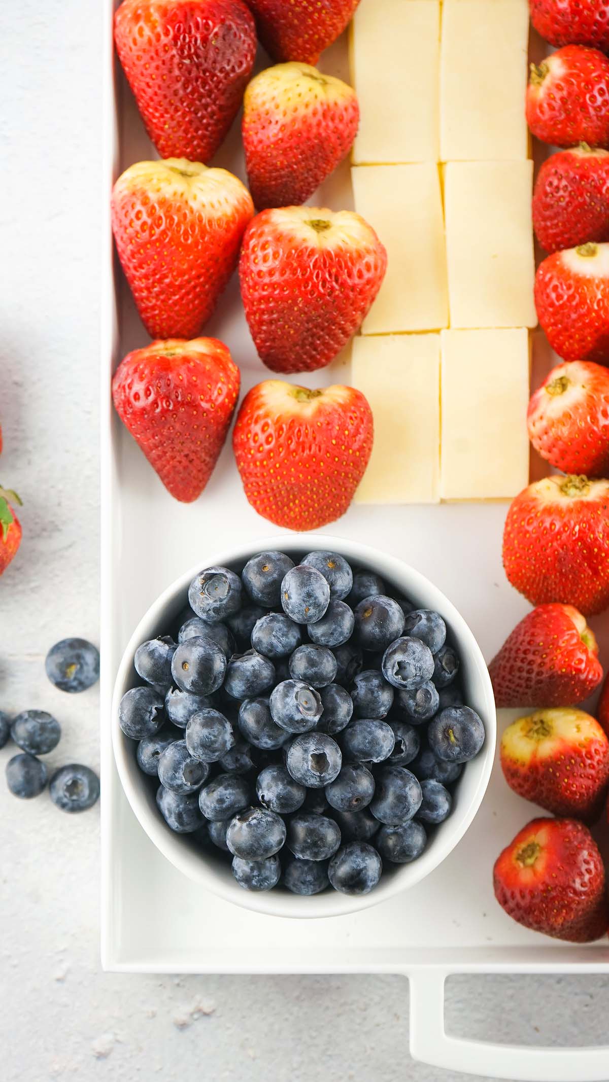 A bowl of blueberries, strawberries, and cheese cubes arranged on a white tray creates a festive Stars and Stripes Cheese Board.