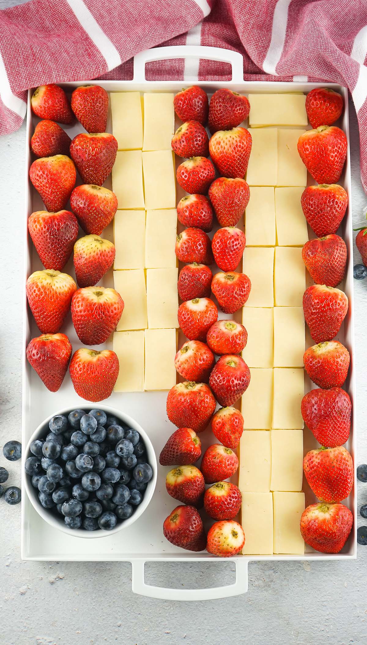 A Stars and Stripes Cheese Board features rows of strawberries and cheese cubes, plus a bowl of blueberries, all arranged neatly on a white surface.