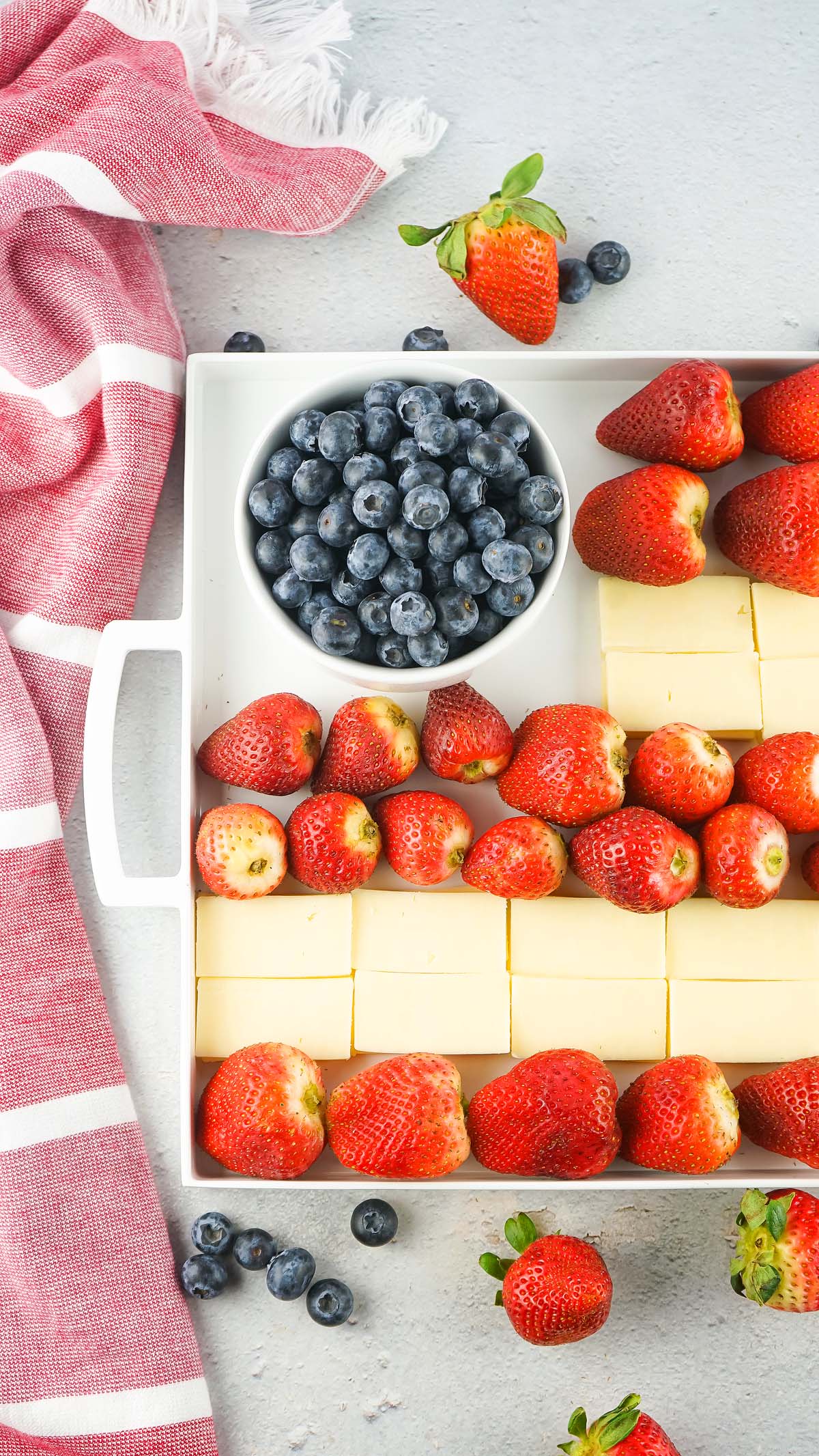Stars and Stripes Cheese Board: White tray with strawberries, blueberries in a bowl, and cheese slices, next to a red striped cloth.