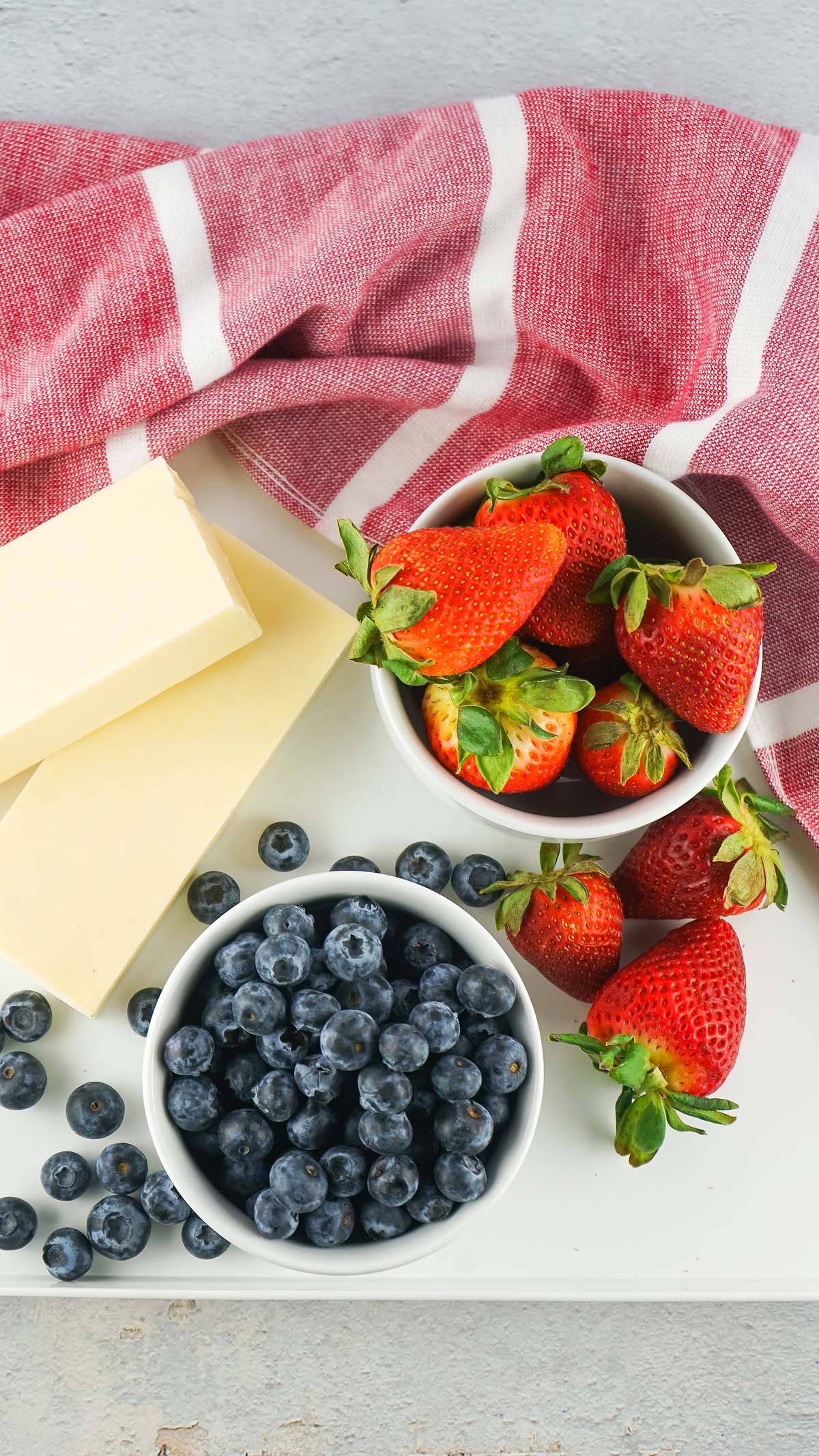 Strawberries, blueberries, and two sticks of butter are arranged on a white surface with a red striped cloth, perfect for assembling your festive Stars and Stripes Cheese Board.