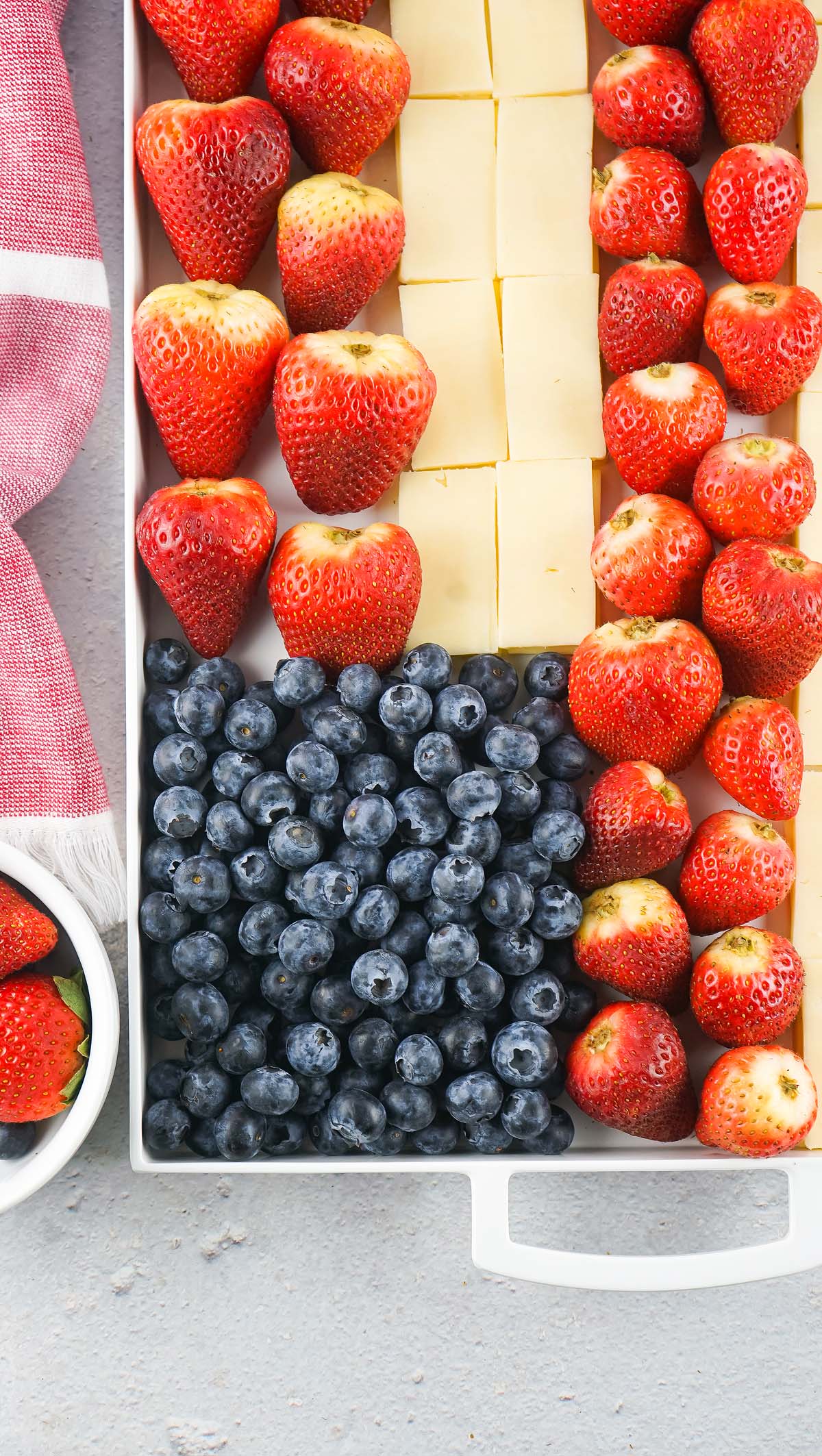 Rectangular tray with rows of strawberries, cheese cubes, and blueberries arranged like a Stars and Stripes Cheese Board, next to a red napkin.