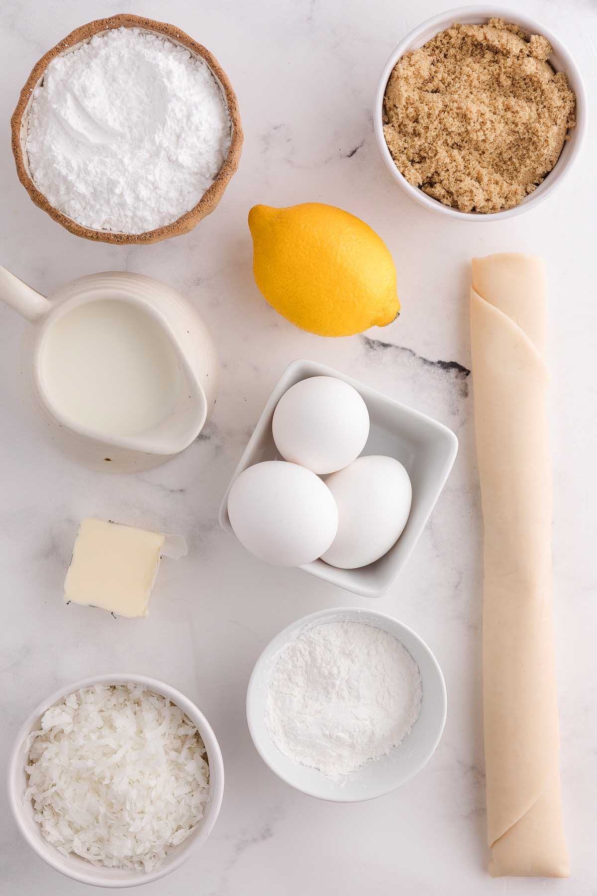 Baking ingredients for coconut meringue pie, including eggs, lemon, milk, butter, sugar, coconut, flour, and rolled dough on a marble surface.