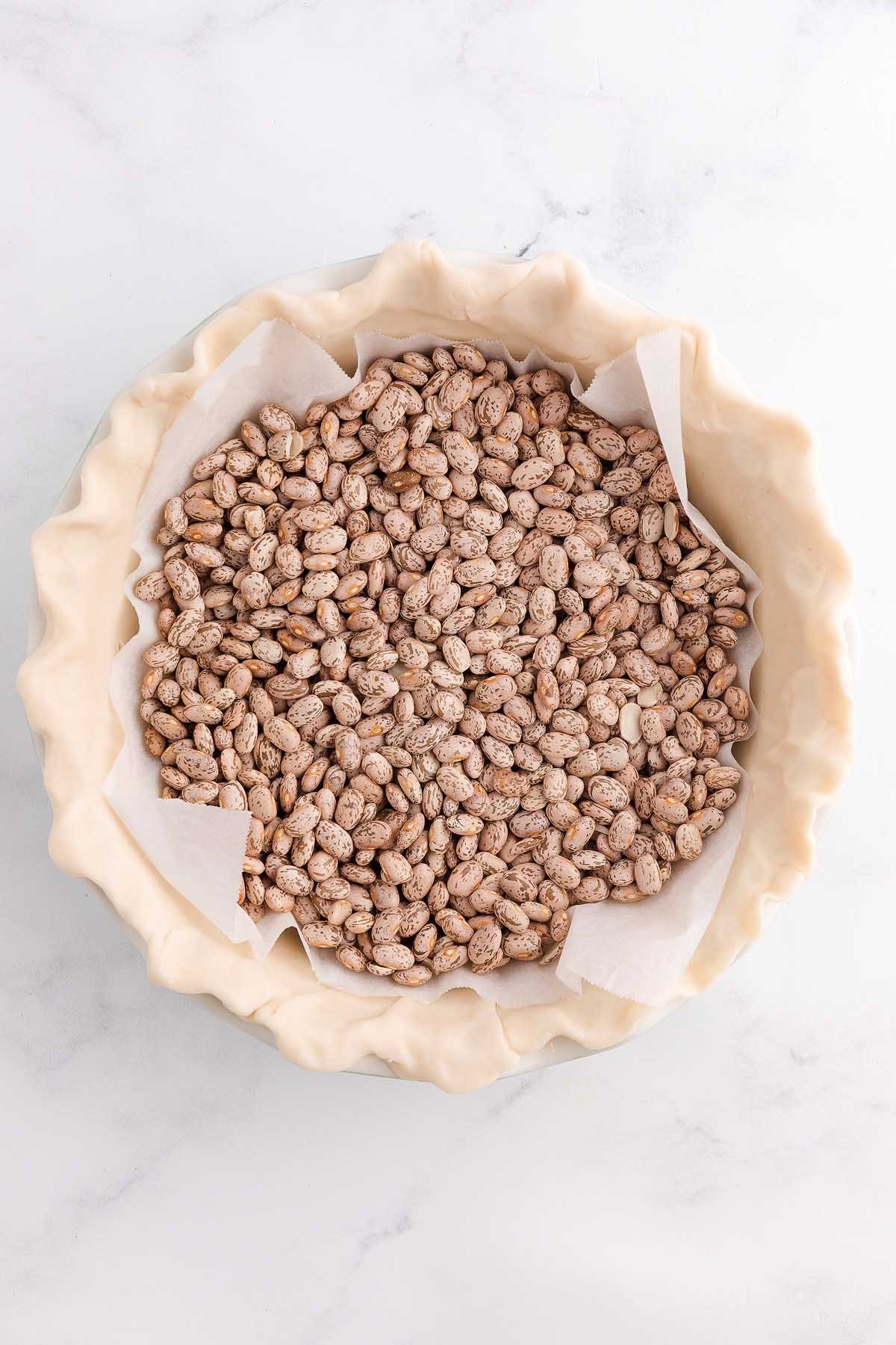 Unbaked pie crust in a pie dish filled with dried beans on parchment paper for blind baking, perfect as a base for a classic coconut meringue pie.