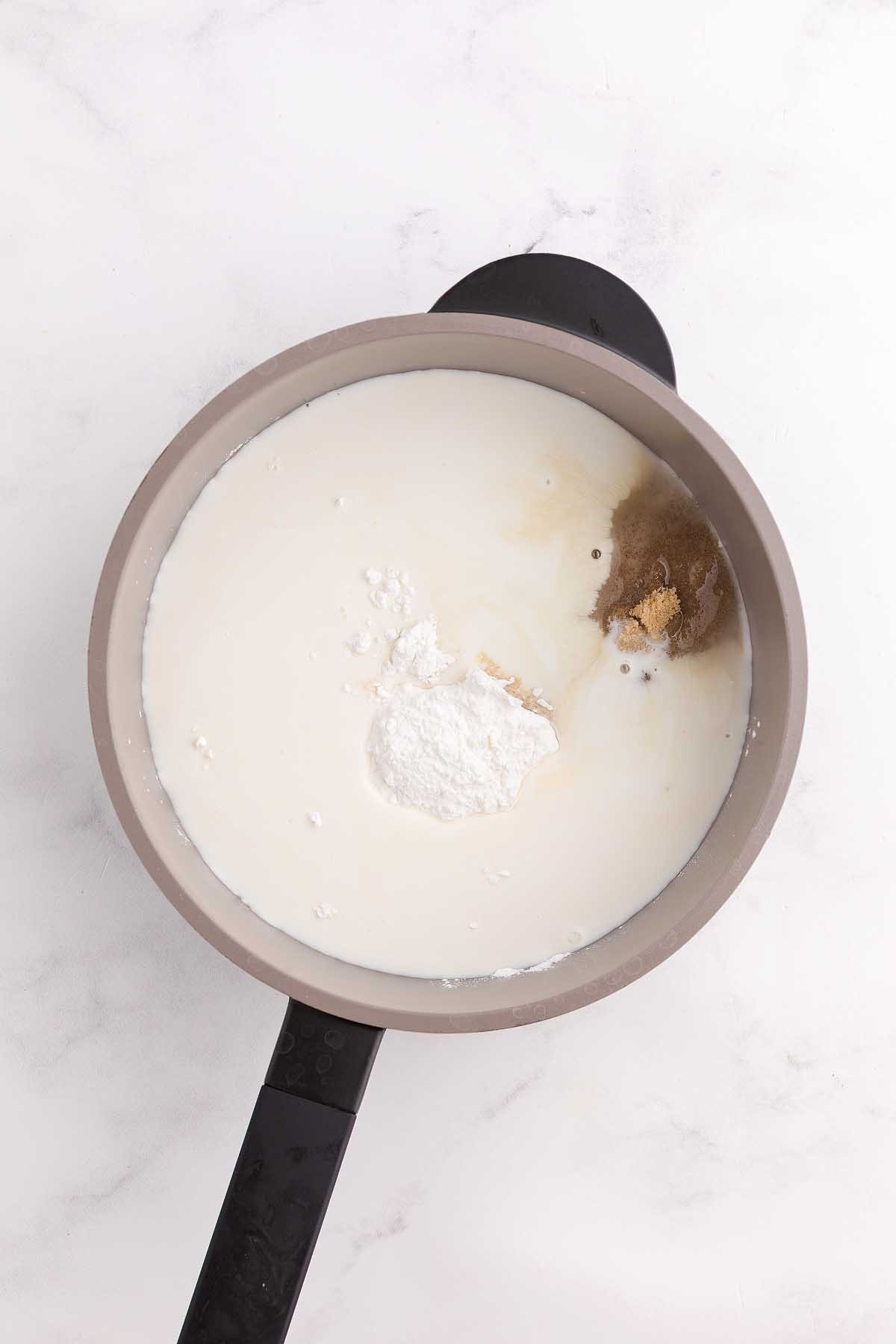A saucepan with milk, brown sugar, cornstarch, and spices on a white marble surface—perfect for starting a classic coconut meringue pie.