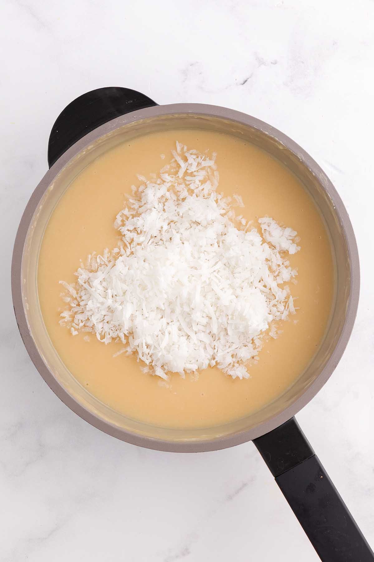 A saucepan filled with a creamy coconut meringue pie mixture, topped with shredded coconut, on a white surface.