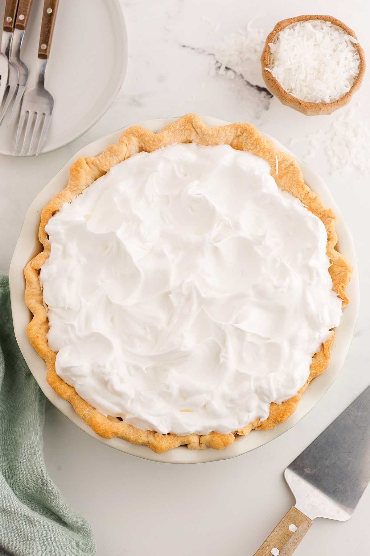 A coconut meringue pie topped with fluffy whipped cream sits on a marble surface, with plates and shredded coconut nearby.