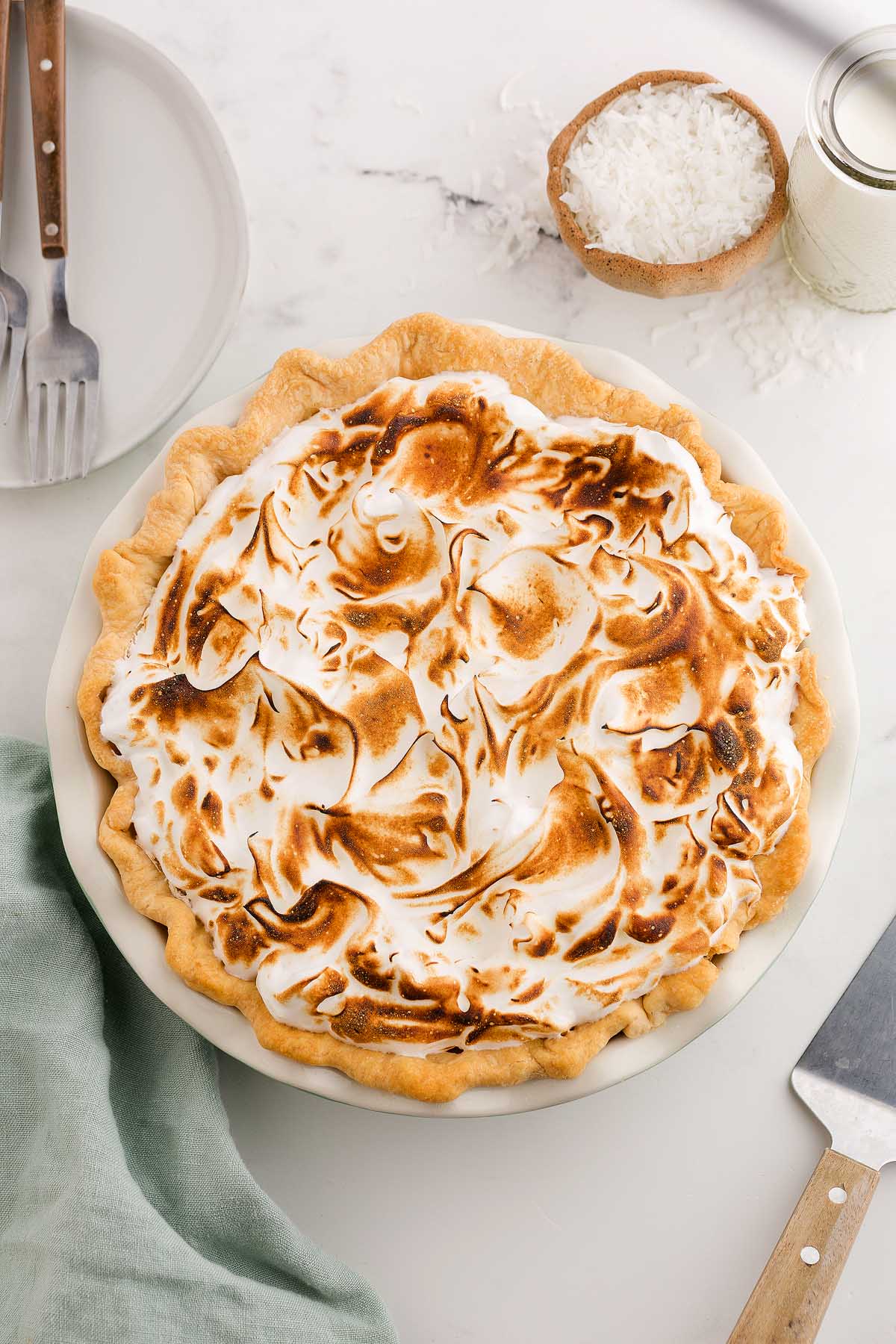 A toasted coconut meringue pie with swirled meringue on top, set on a white table alongside plates and utensils.