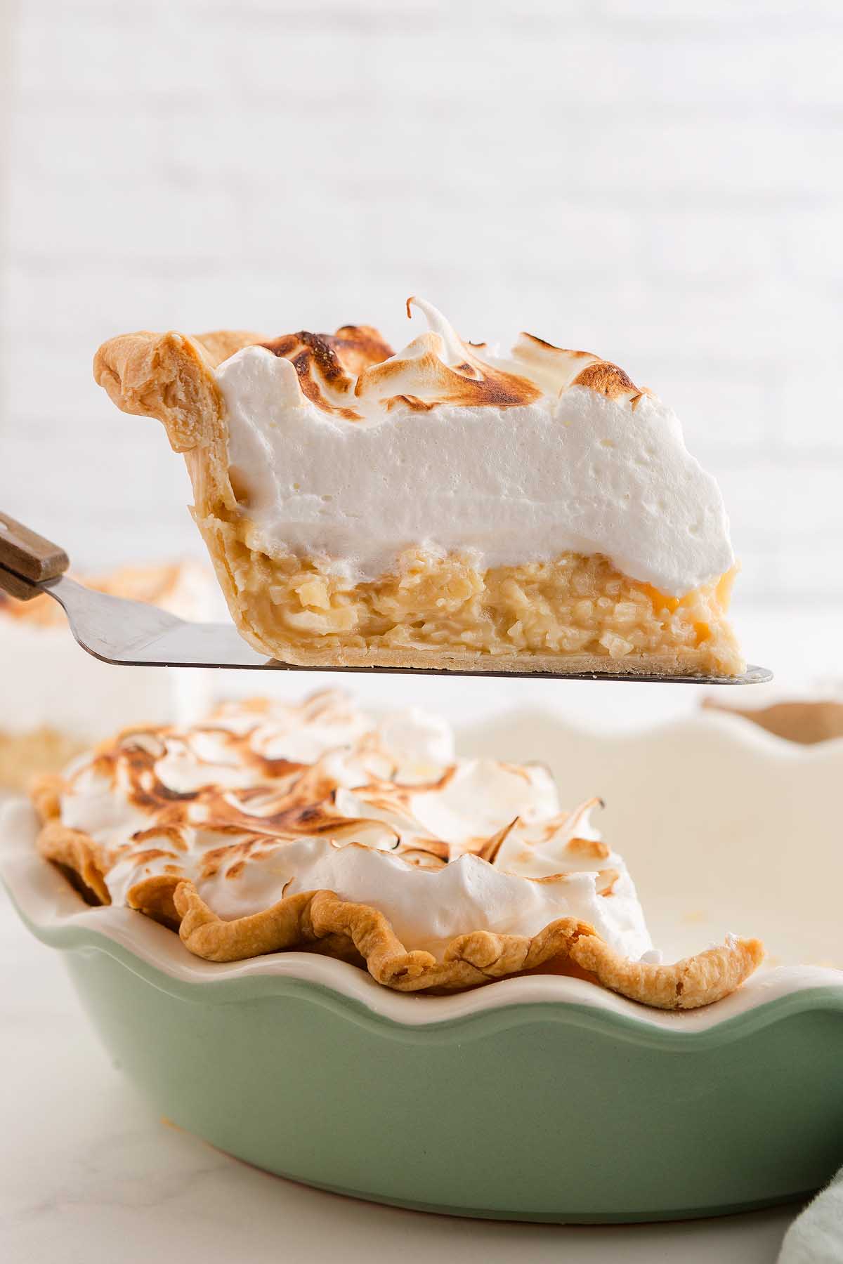 A slice of coconut meringue pie, crowned with golden peaks, is being lifted from a green pie dish.