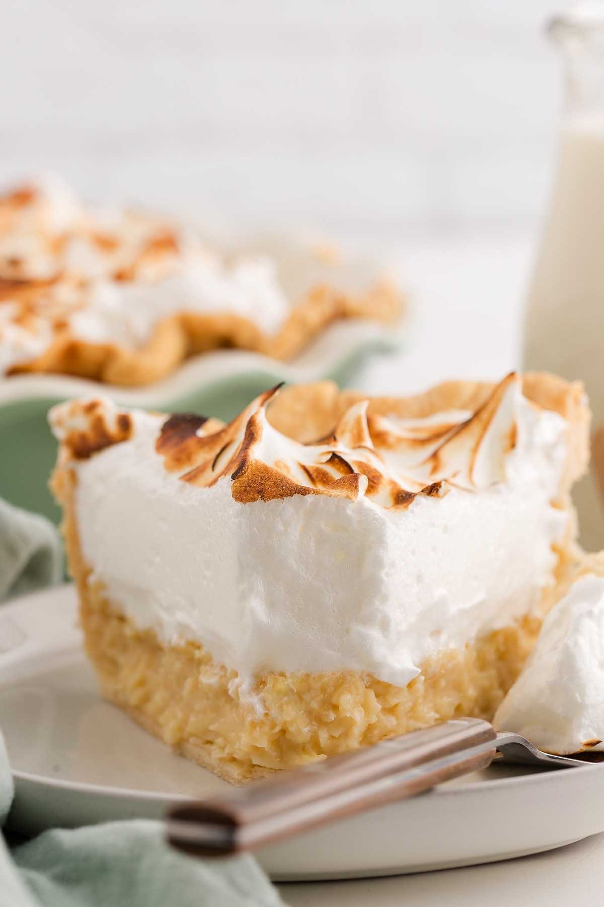 A slice of coconut meringue pie with toasted meringue topping sits on a plate, a fork beside it.