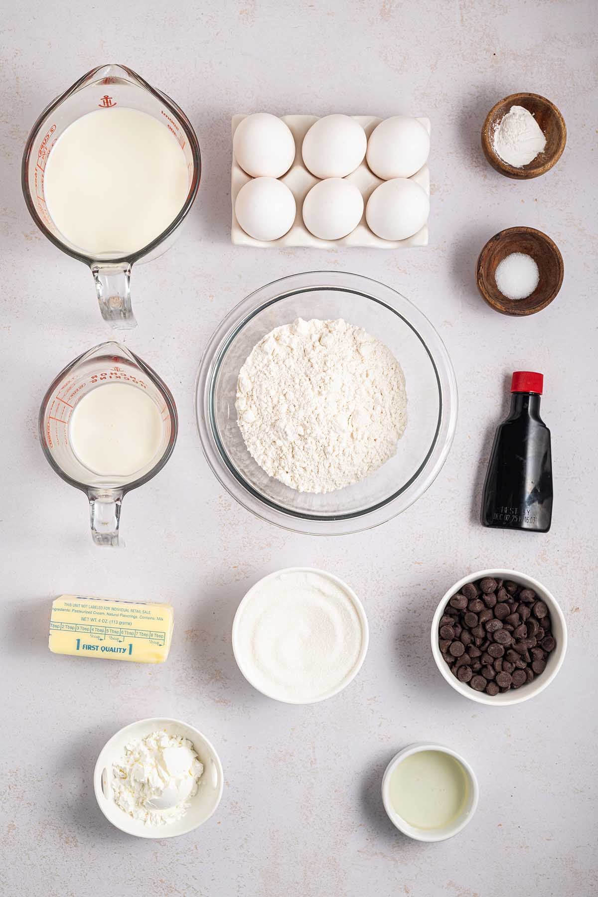 Overhead view of baking ingredients for Boston cream pie: eggs, flour, sugar, milk, butter, chocolate chips, and vanilla extract.
