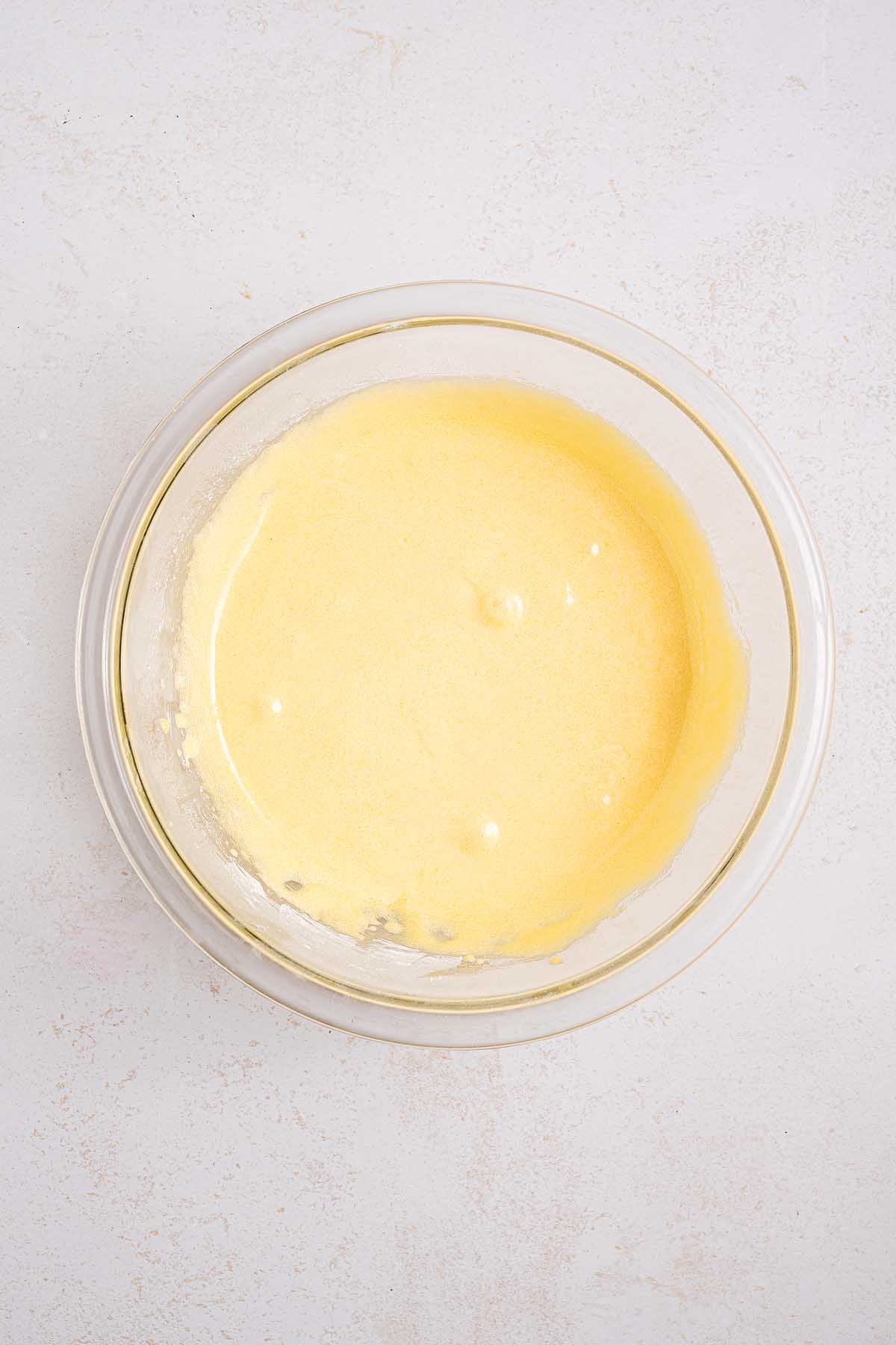 A glass bowl filled with smooth, yellow Boston cream pie batter rests on a light-colored surface.