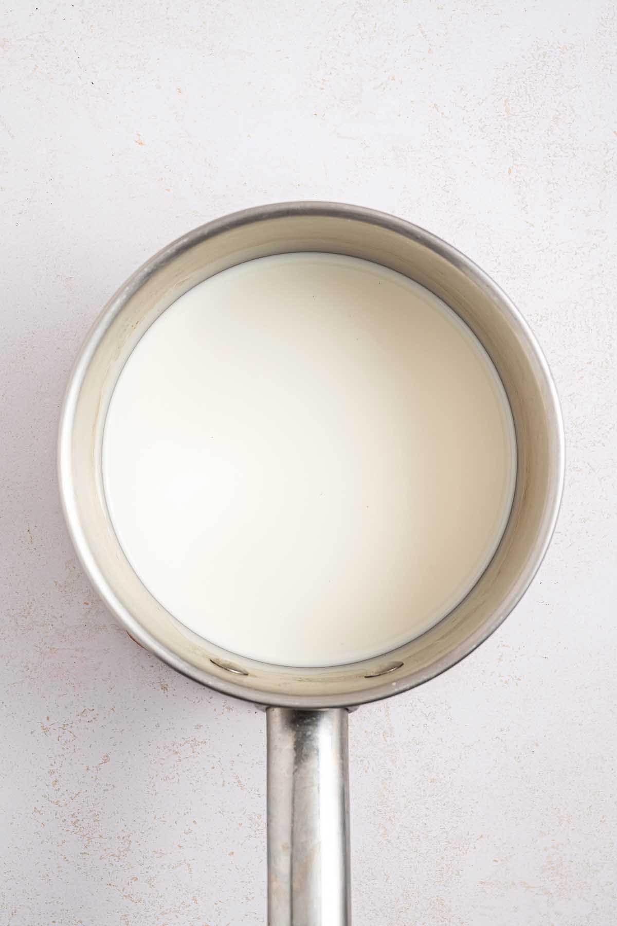 A metal saucepan filled with milk, perfect for starting a classic Boston cream pie, sits on a light-colored surface, viewed from above.