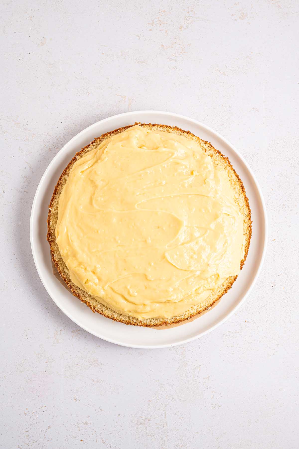 A round Boston cream pie with yellow frosting sits on a white plate, photographed from above on a light surface.