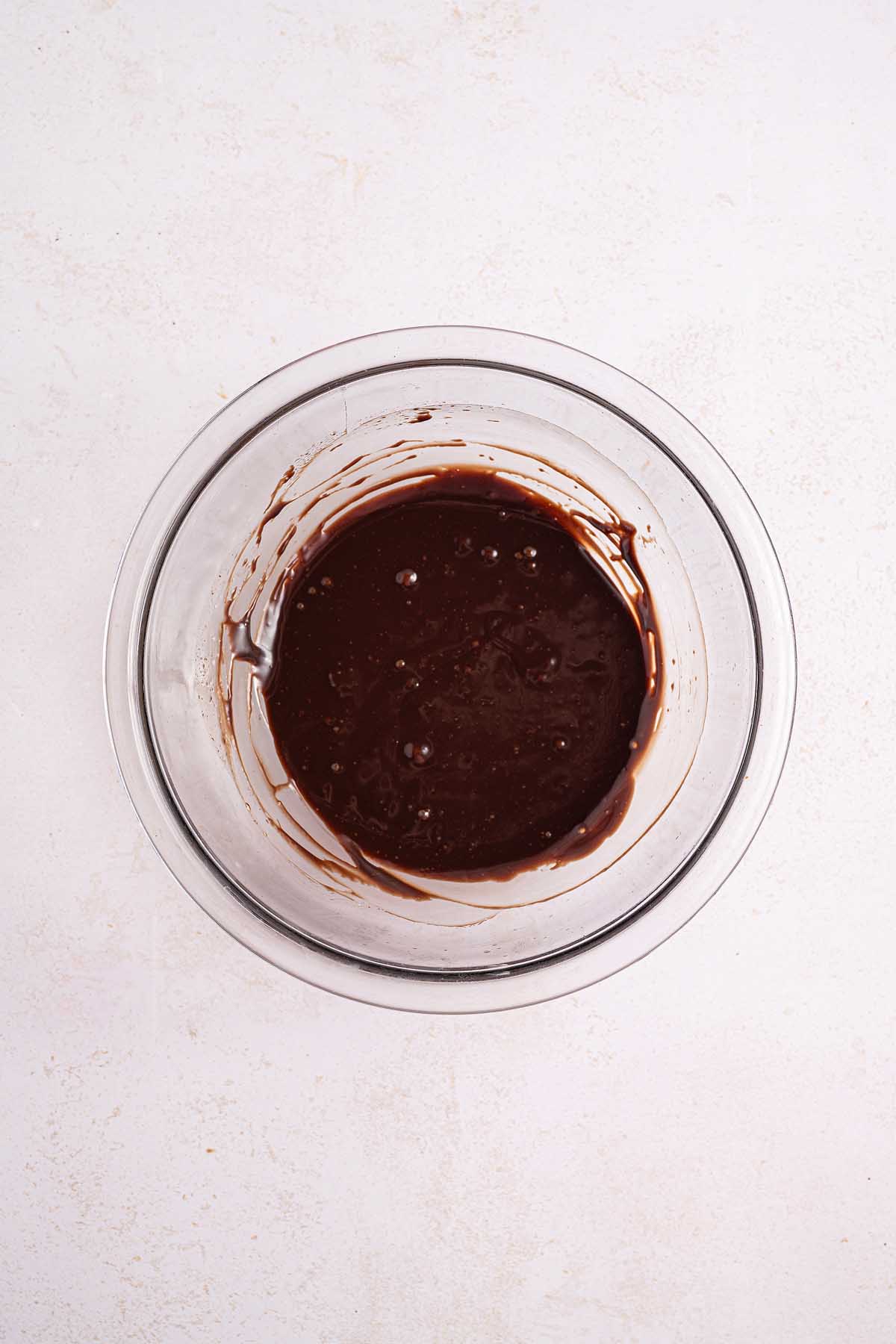 A glass bowl filled with rich melted chocolate mixture, perfect for topping a classic Boston cream pie, sits on a light-colored surface.