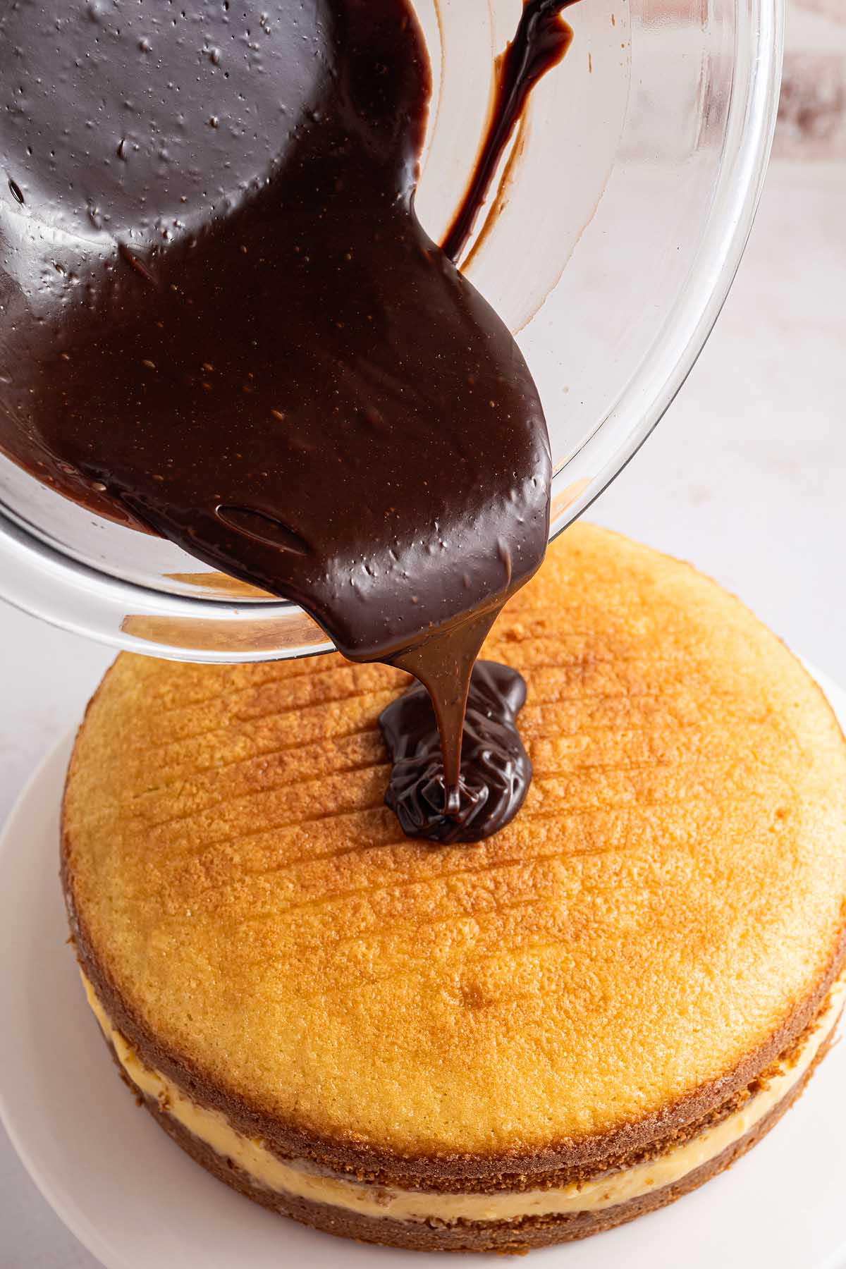 Chocolate ganache being poured from a bowl onto a layered yellow cake on a white plate, creating a classic Boston cream pie look.