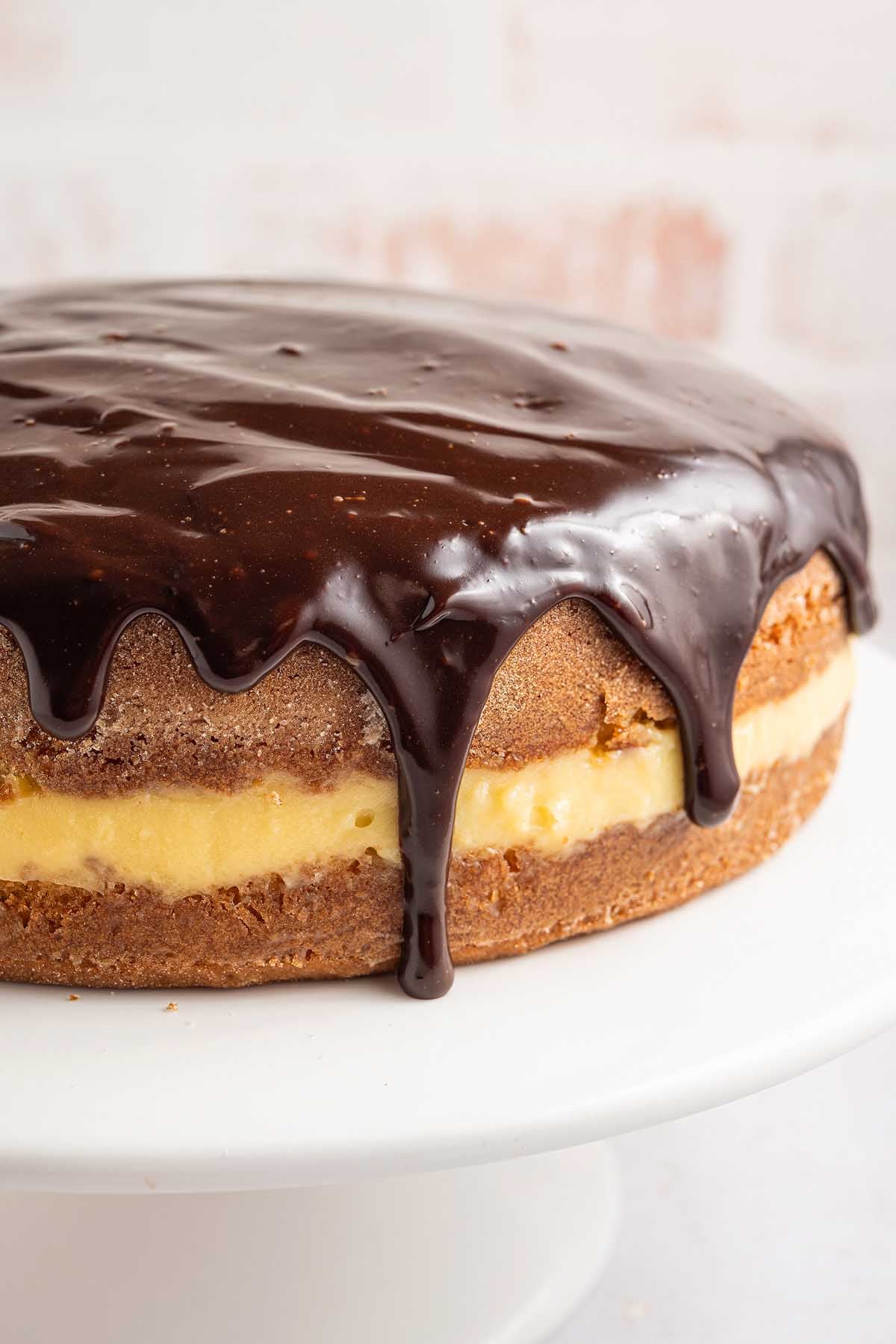 Boston cream pie with rich chocolate ganache dripping over silky yellow custard filling, beautifully presented on a white cake stand.