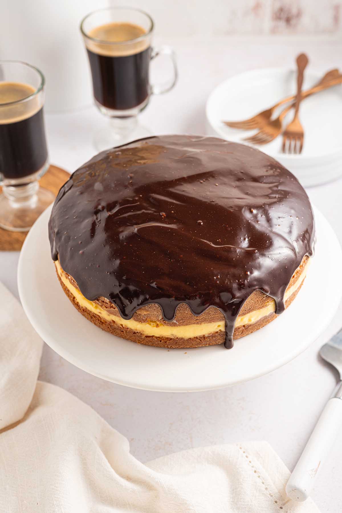 A round Boston cream pie with glossy chocolate glaze sits on a white stand, next to two cups of coffee and plates with forks.