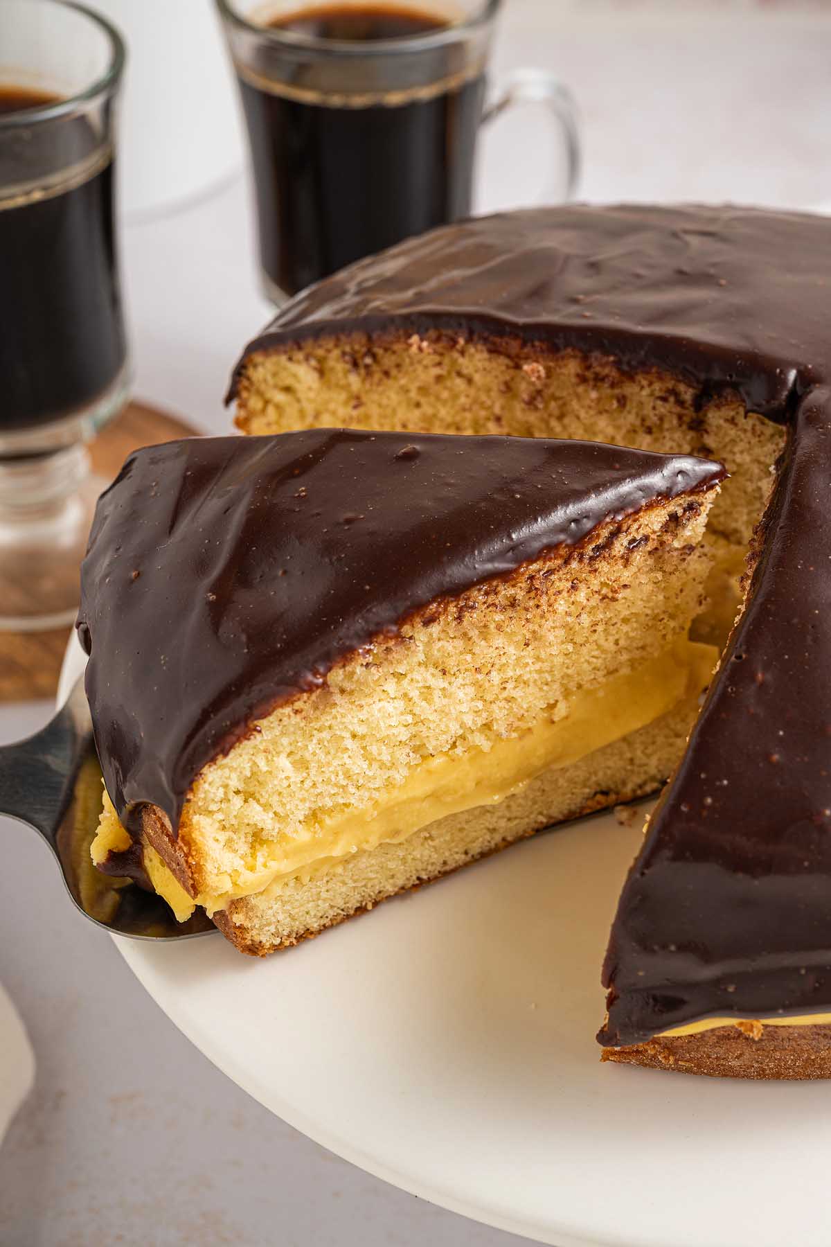 A slice of Boston cream pie being lifted from the decadent cake, with two cups of coffee in the background.