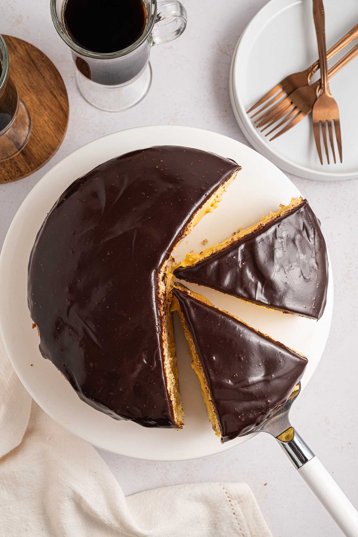 A Boston cream pie-inspired chocolate-glazed cake with two slices cut, served on a white plate alongside coffee and forks.