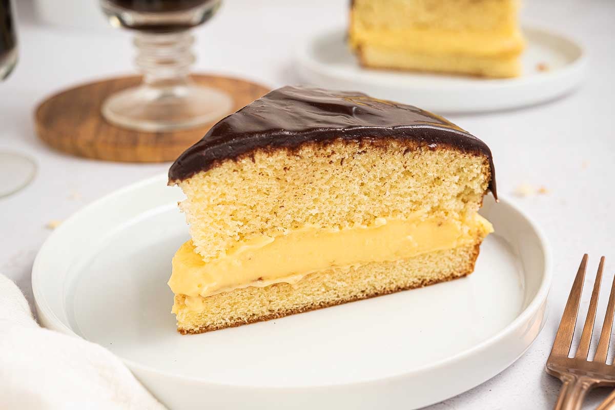 A delicious slice of Boston cream pie, featuring rich chocolate glaze and creamy custard filling, sits on a white plate.