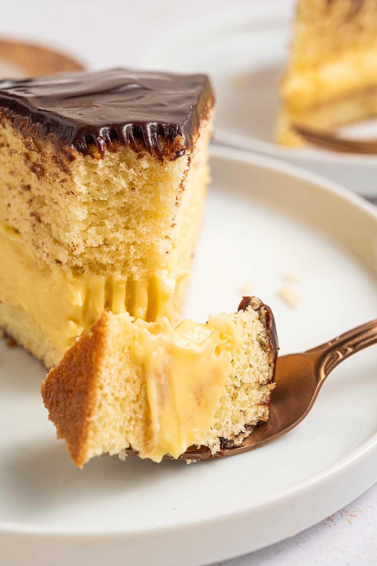 A slice of Boston cream pie sits on a white plate, with a bite taken on a fork, showcasing its creamy filling and rich chocolate glaze.