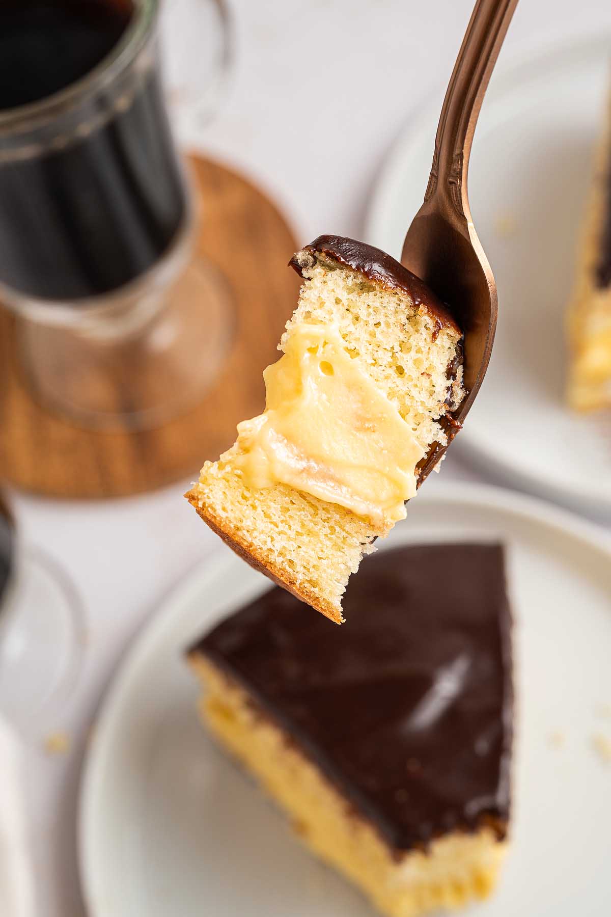A fork holds a bite of classic Boston cream pie, showcasing its rich custard filling and glossy chocolate glaze above a plate.