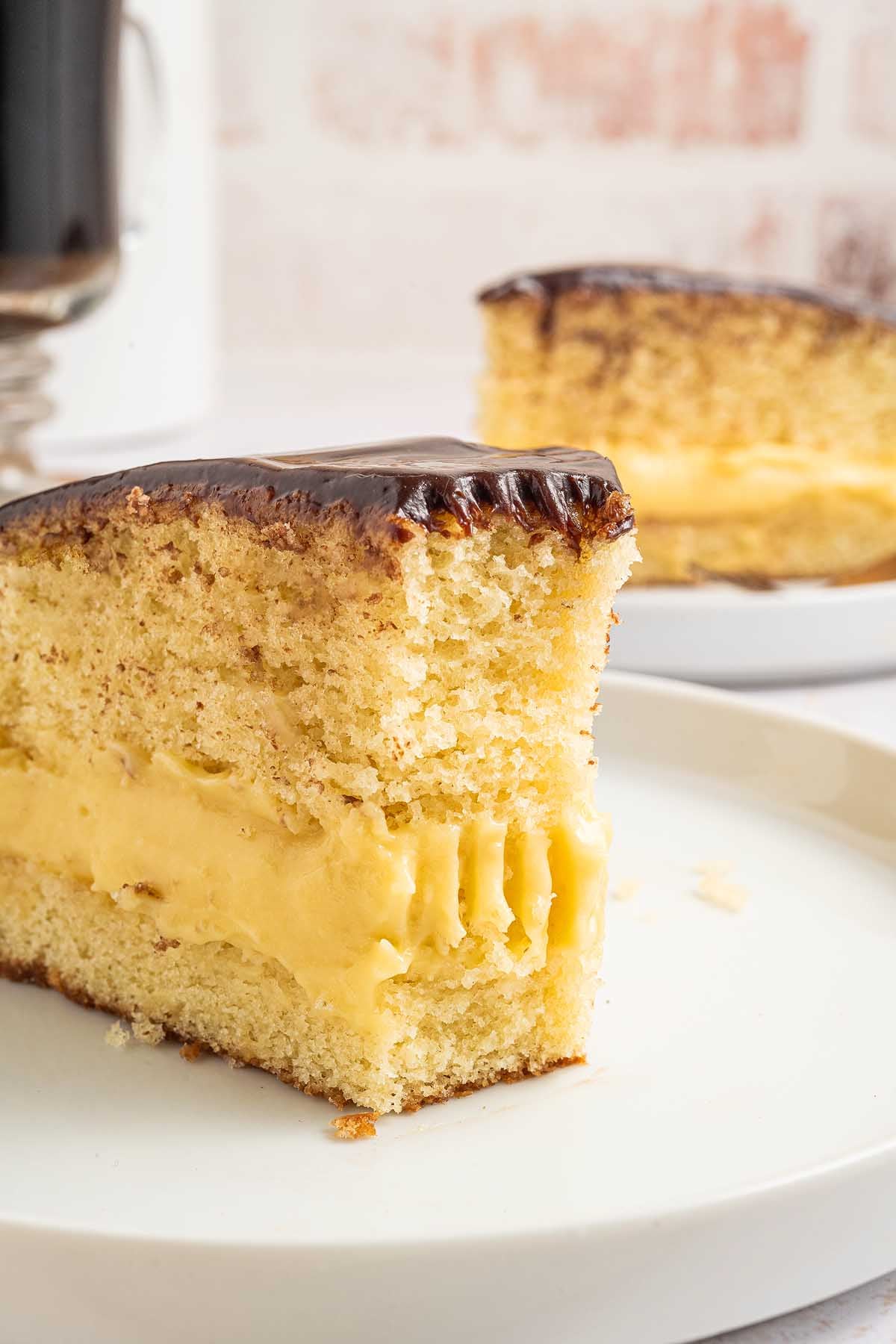 A slice of Boston cream pie with rich chocolate glaze and creamy custard filling sits on a white plate.