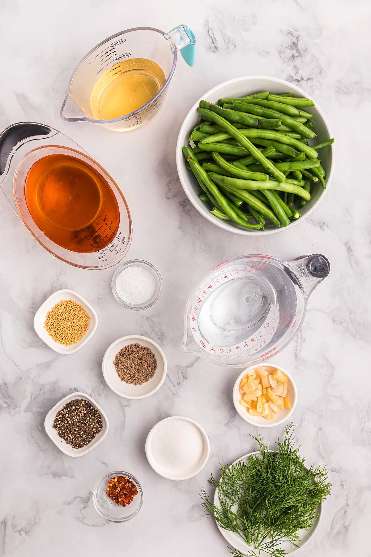 Fresh pickled green beans, liquids, herbs, and spices arranged in bowls and pitchers on a marble countertop.