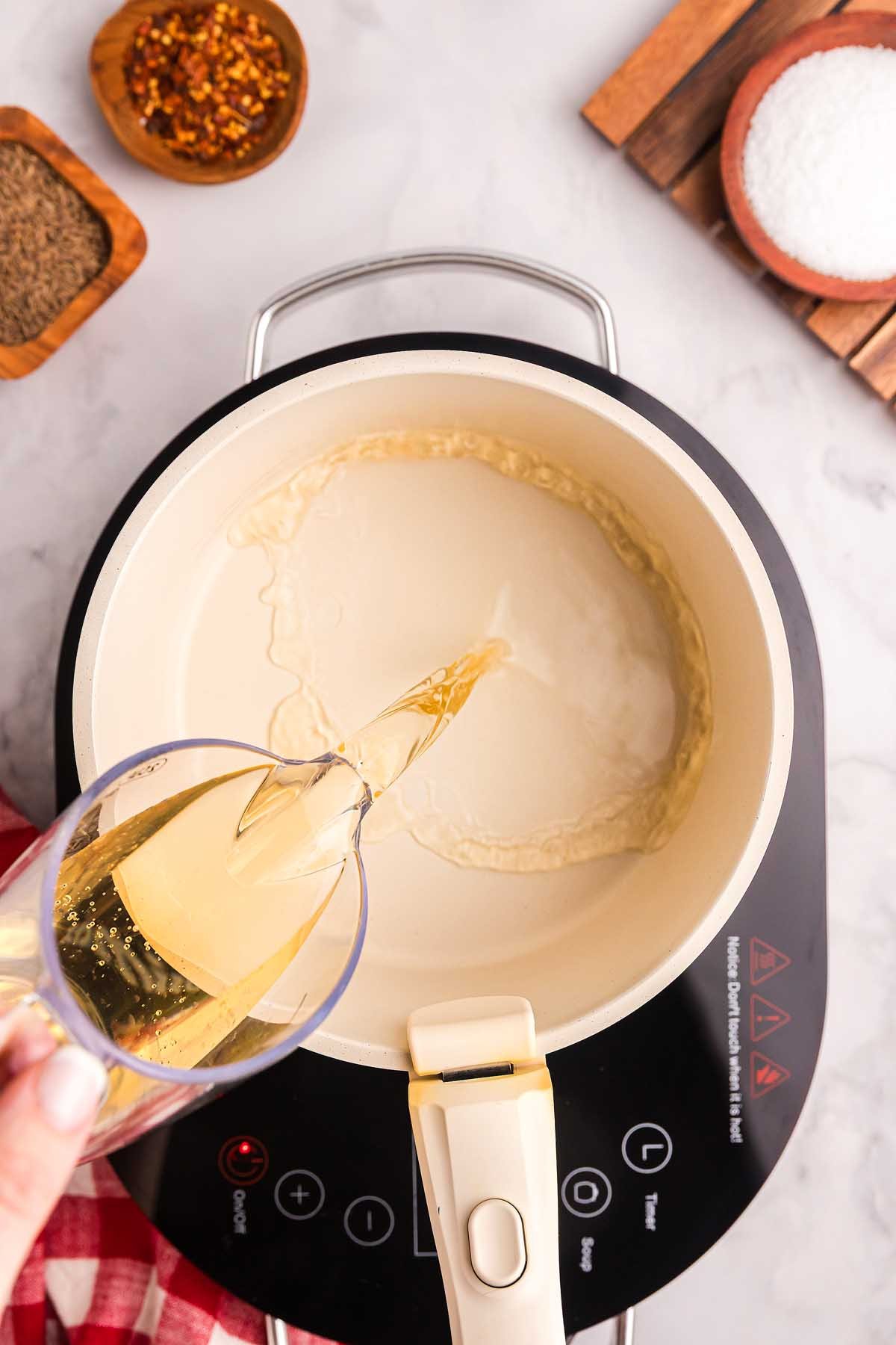 A hand pours ingredients from a glass cup into a white pot on an electric stove, with spices and pickled green beans nearby.