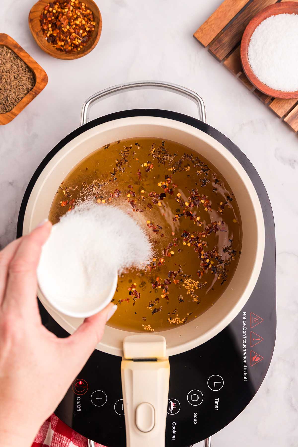 Hand pouring ingredients into a pot with spices on an electric cooktop, surrounded by bowls of ingredients for making pickled green beans.