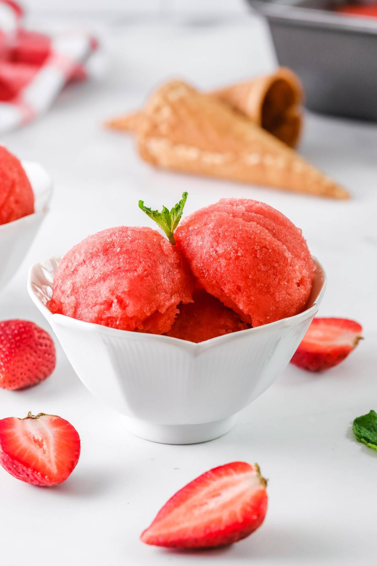 White bowl with two scoops of refreshing strawberry sorbet, made with frozen strawberries and just 4 ingredients, garnished with mint and surrounded by fresh strawberries.
