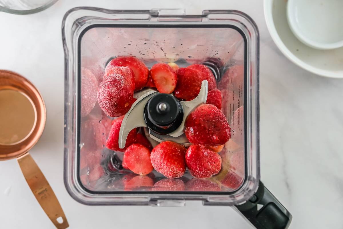 Frozen strawberries in a blender, viewed from above, with a measuring cup and plates nearby on a white surface—perfect for a summer dessert idea like sorbet with just 4 ingredients.