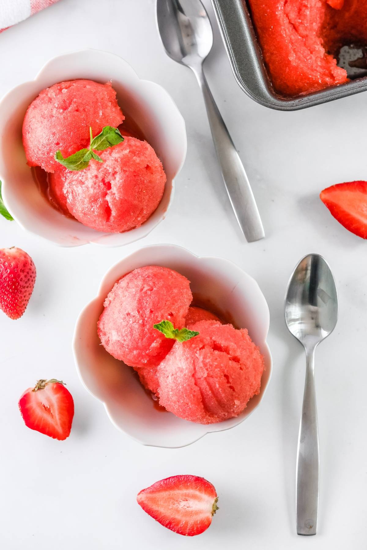 Two white bowls with refreshing strawberry sorbet made with just 4 ingredients, garnished with mint and surrounded by fresh strawberries and spoons.