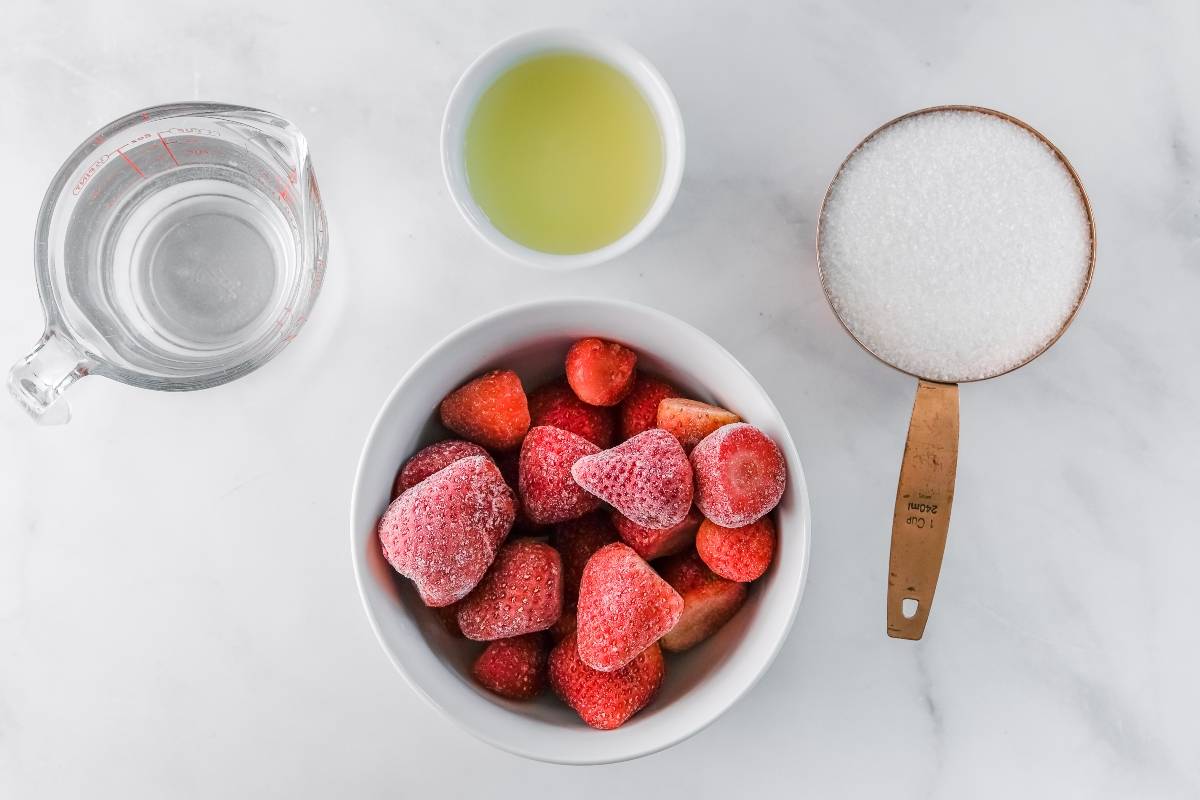Refreshing Strawberry Sorbet: Frozen strawberries in a bowl, sugar in a measuring cup, lime juice, and water on a white surface for a quick and easy sorbet.