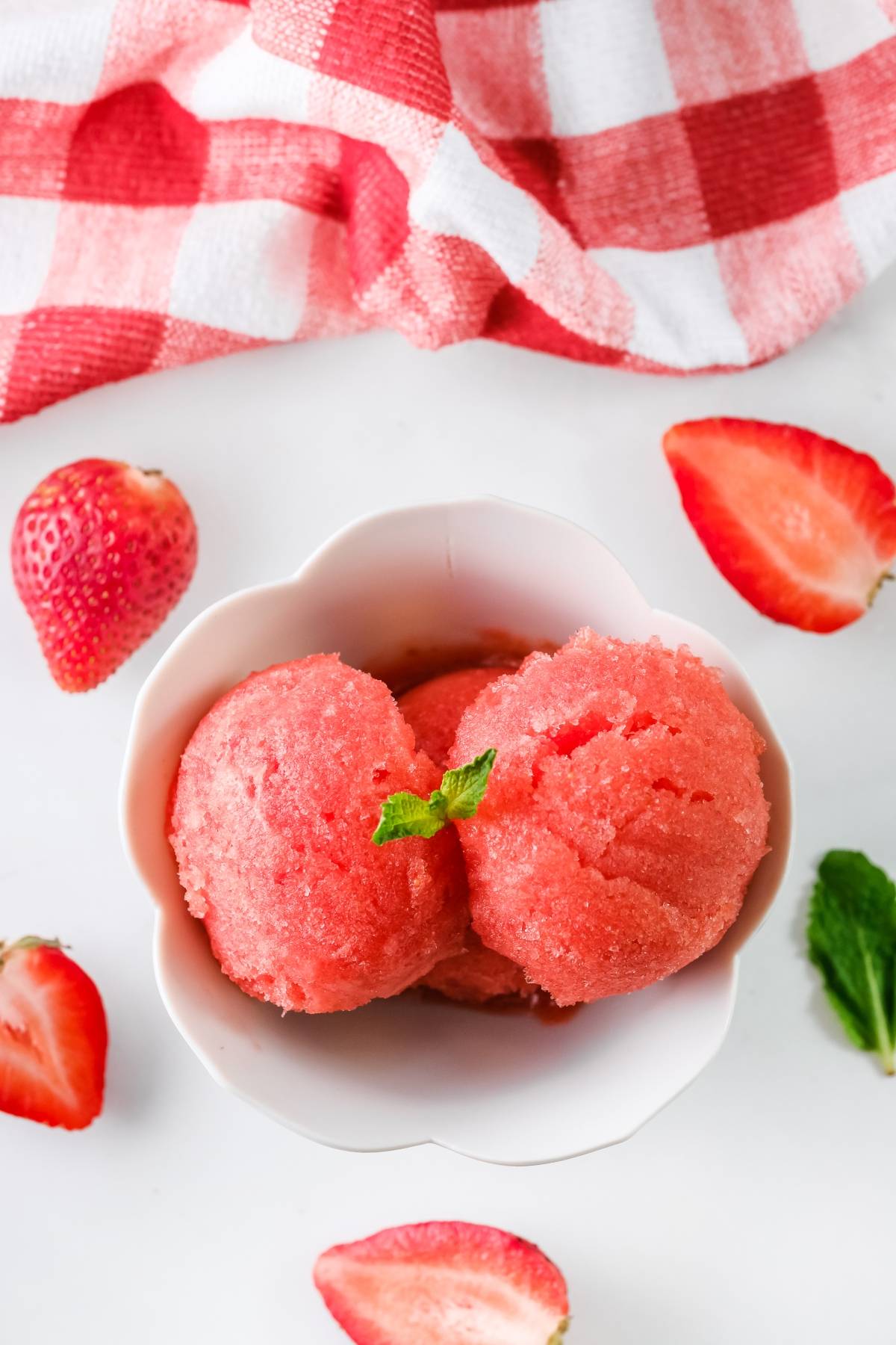 Two scoops of strawberry sorbet in a white bowl, garnished with mint, beside fresh strawberries and a red towel. Made with frozen strawberries, it's the perfect summer dessert idea for a refreshing treat.