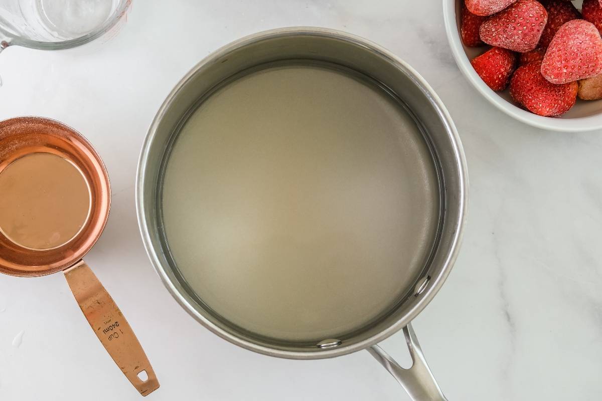 A pot of clear liquid, a copper measuring cup, and a bowl of strawberries on a white surface—perfect essentials for making quick and easy sorbet or refreshing strawberry sorbet made with frozen strawberries.