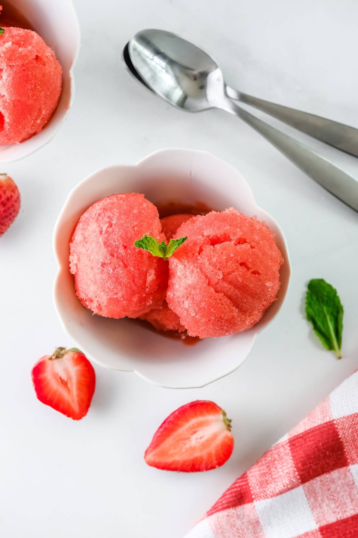 Two scoops of refreshing strawberry sorbet, made with frozen strawberries, nestled in a white bowl and garnished with mint—sliced strawberries and spoons nearby make this the perfect summer dessert idea.