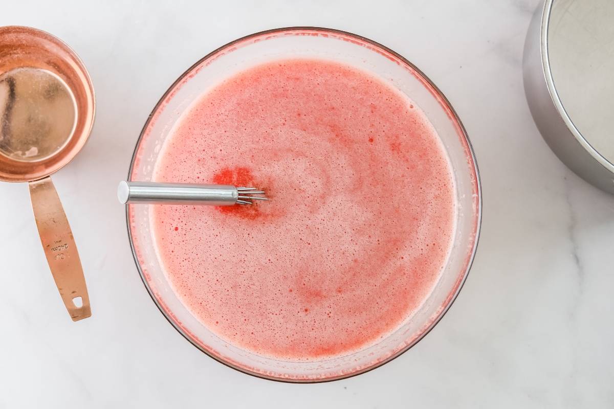 A whisk in a bowl of pink liquid, hinting at a quick and easy sorbet made with frozen strawberries, with a copper measuring cup and metal container nearby on a white surface.