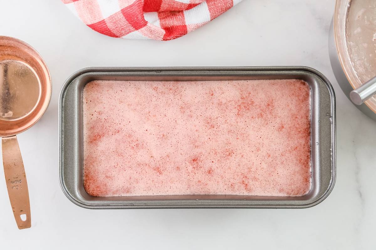 A metal loaf pan filled with a pink, frothy mixture of Quick and easy sorbet sits on a white surface near a copper measuring cup.