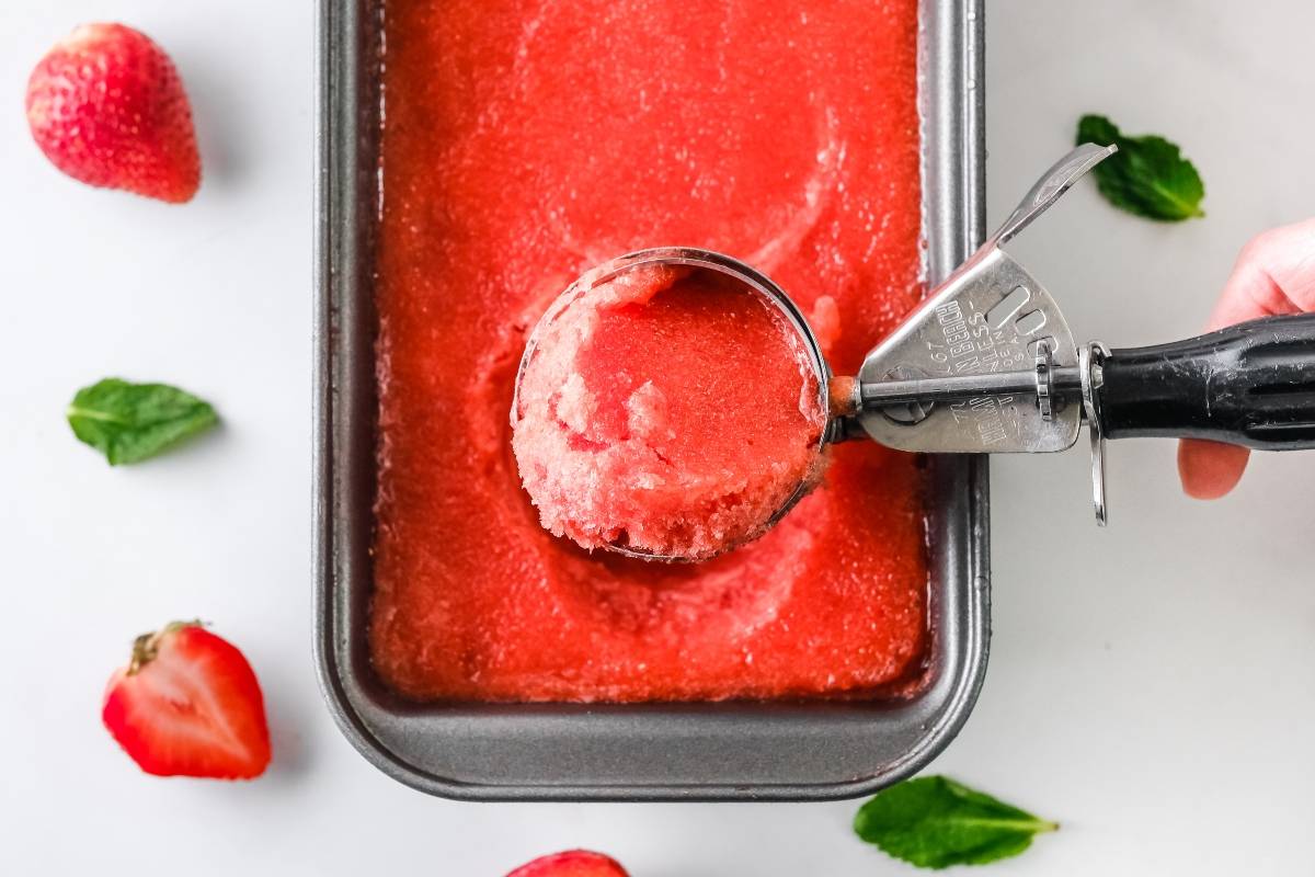 A metal scoop lifts refreshing Strawberry Sorbet from a rectangular pan, surrounded by strawberries and mint leaves. Quick and easy sorbet made with frozen strawberries.