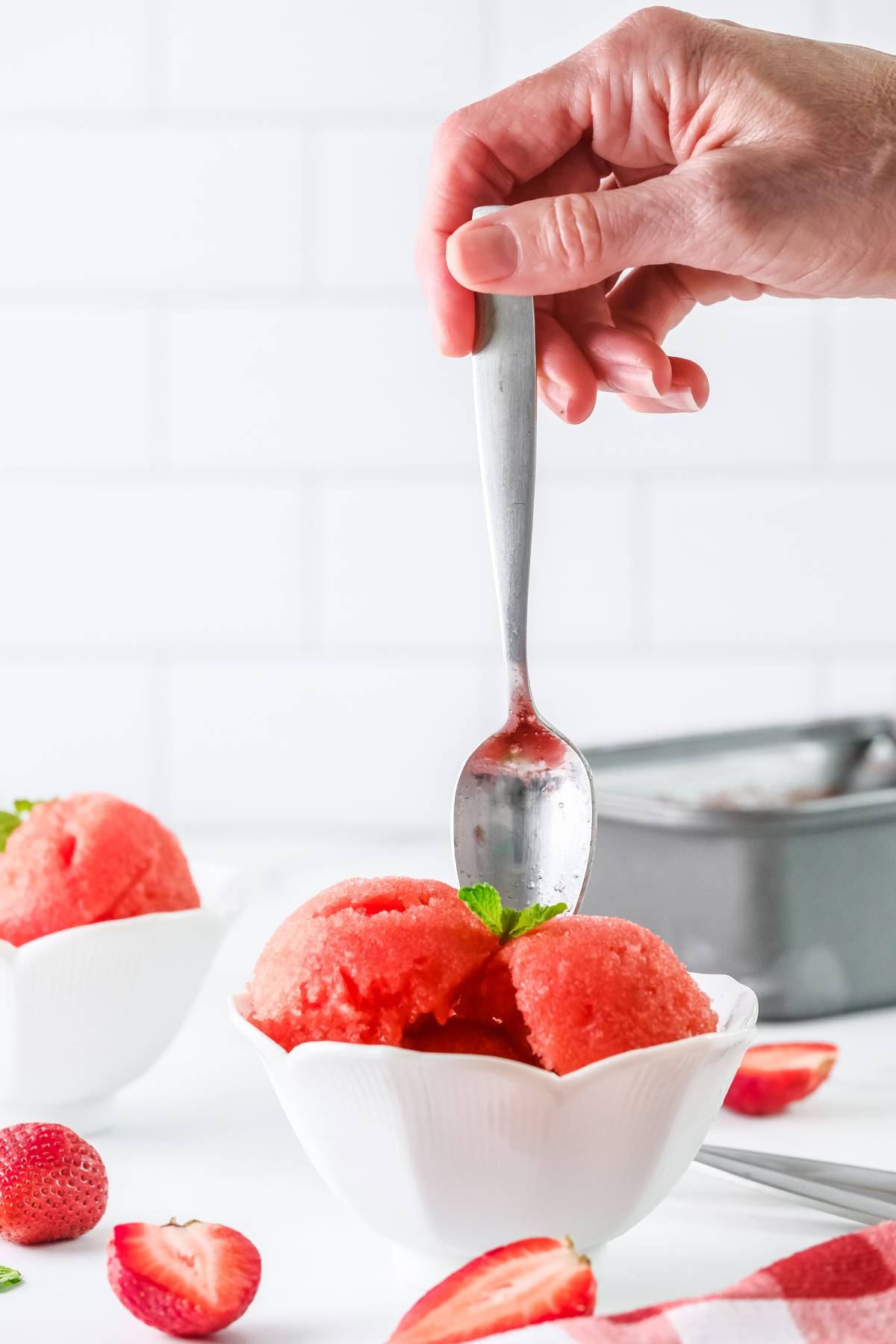 A hand holds a spoon over a bowl of strawberry sorbet made with frozen strawberries, topped with fresh strawberries and mint garnish.