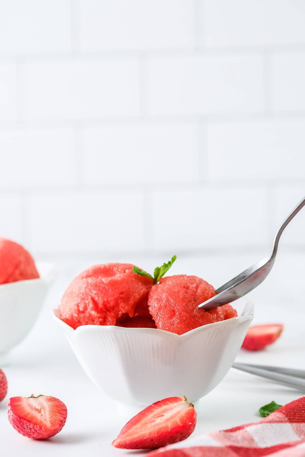 A bowl of refreshing strawberry sorbet with a fork, surrounded by fresh strawberries on a white surface—this sorbet with just 4 ingredients is made with frozen strawberries for the ultimate cool treat.
