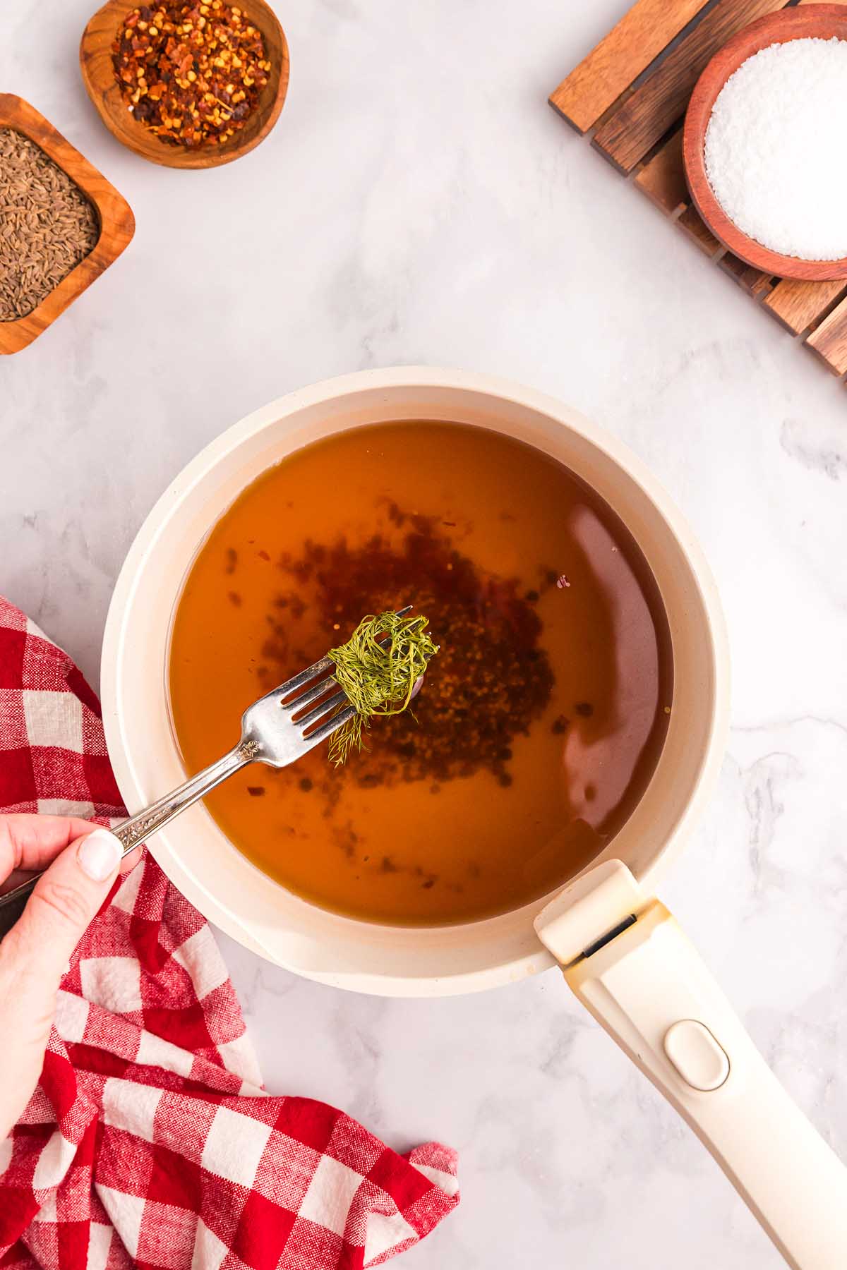 A hand holds a fork with dill over a saucepan of brine for pickled green beans, surrounded by spices and a red checkered towel.